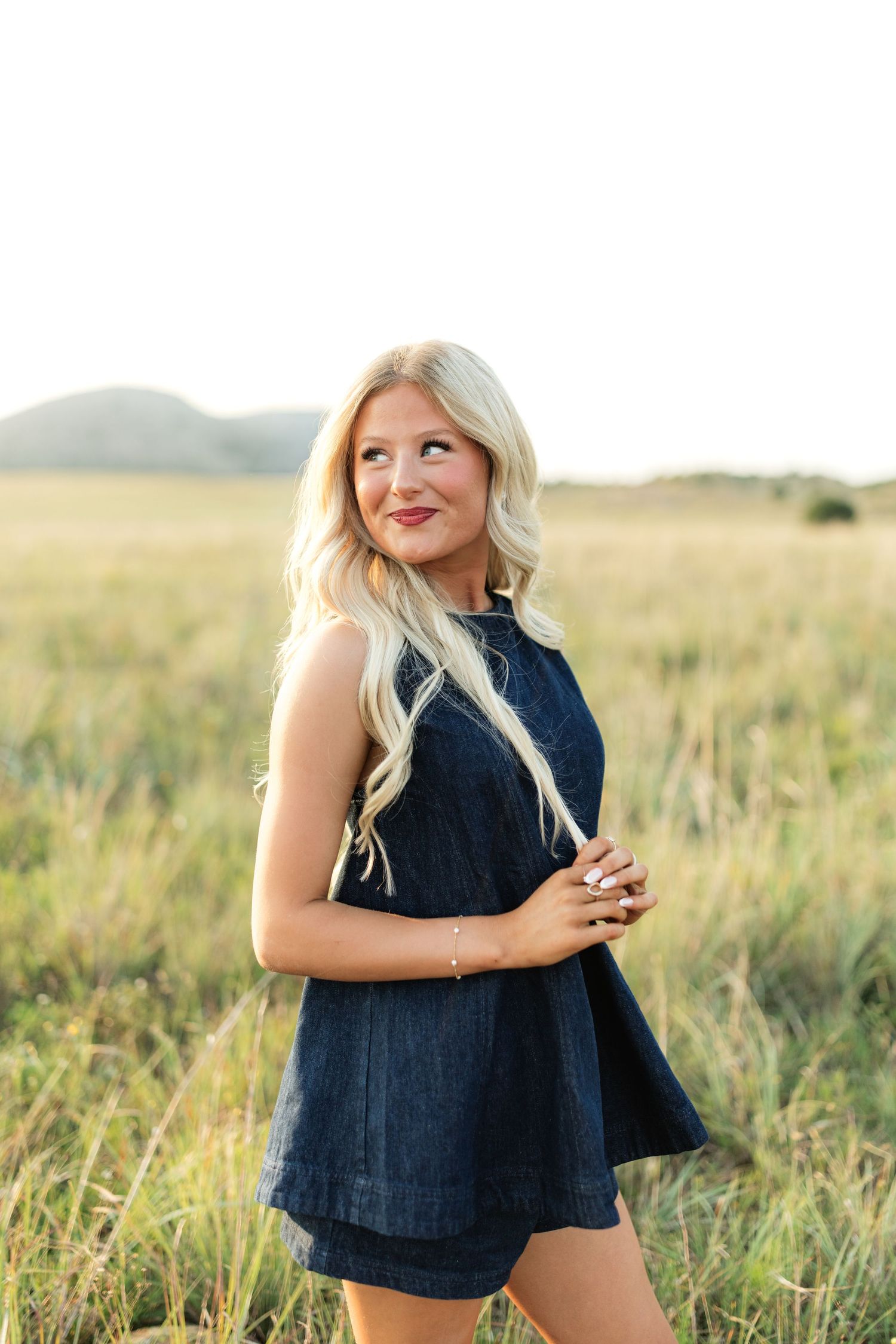 Blonde woman in navy blue sleeveless dress smiling in golden grass field with mountains in background.