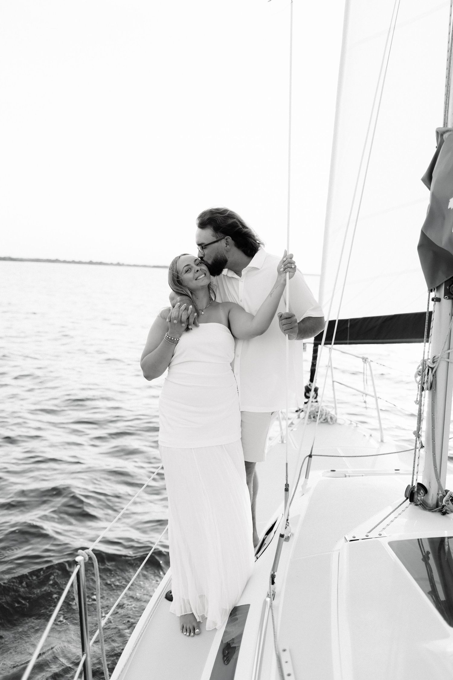 Black and white photo of a couple sharing a romantic kiss on a sailboat deck with calm water in the background.
