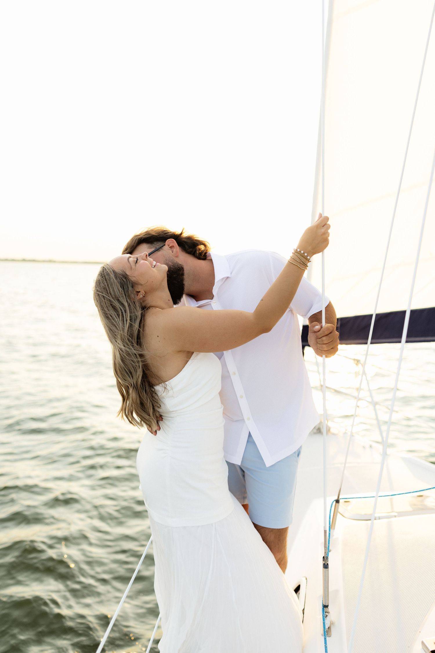 Couple sharing a romantic kiss on a sailboat, dressed in white, with ocean water in the background.