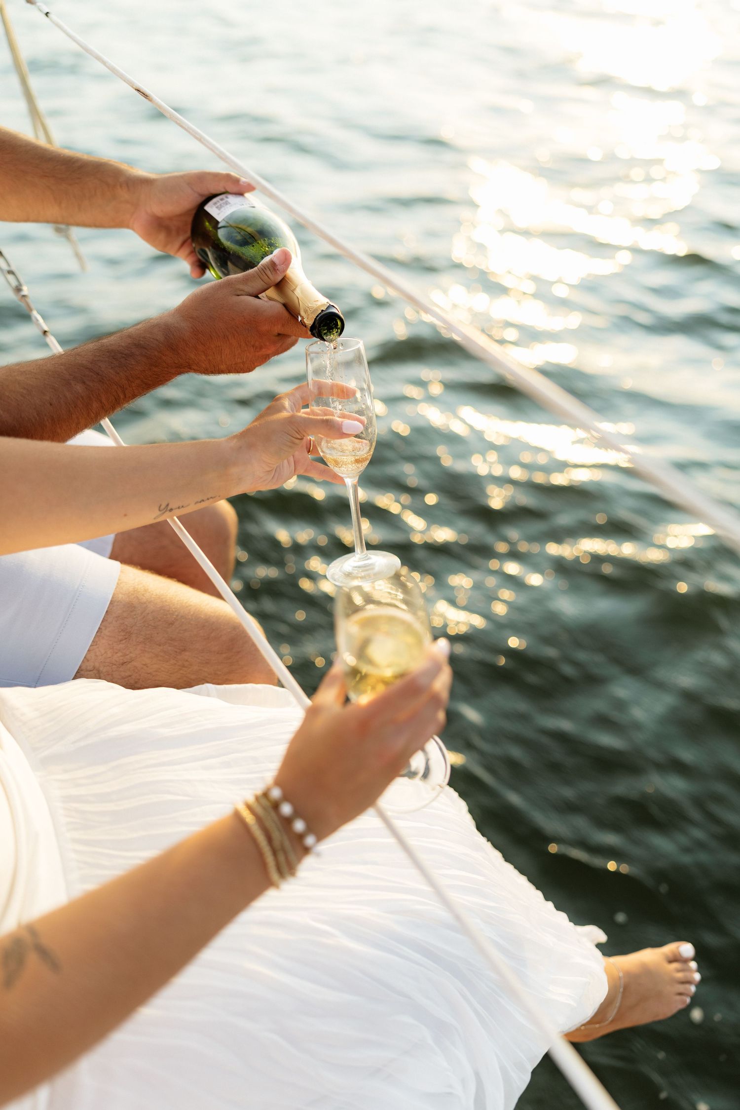 A couple pours champagne into glasses on a sailboat deck with sunlit sparkling water in the background.