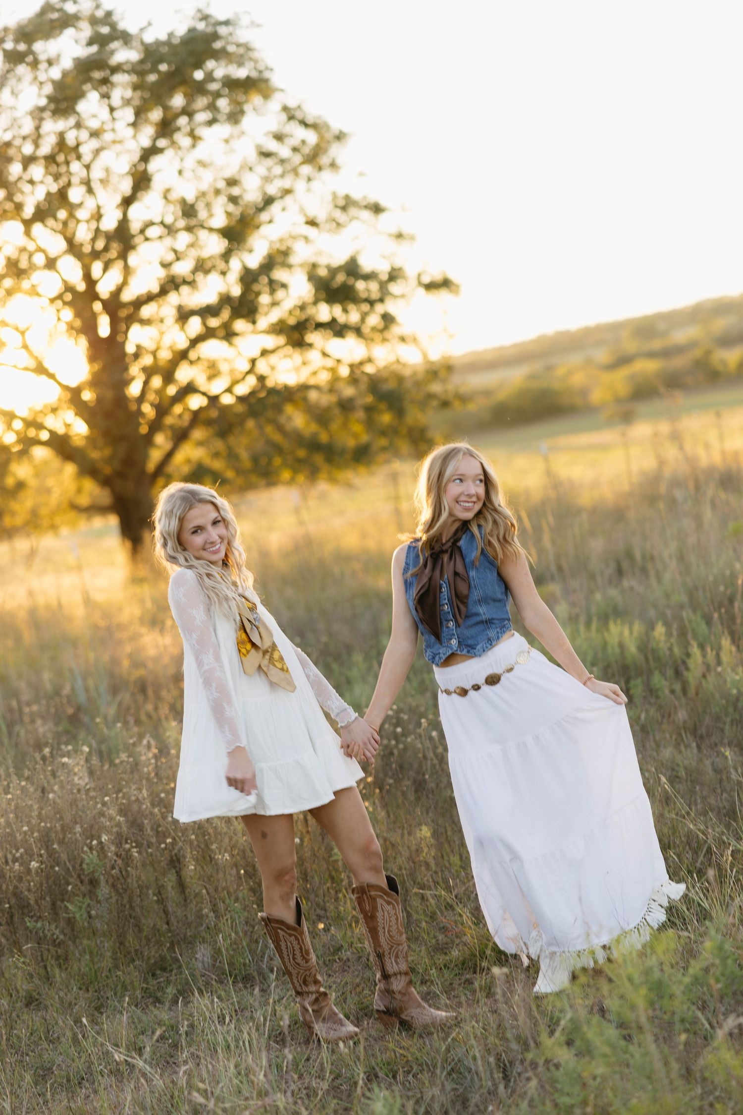 Two smiling women in white dresses and cowboy boots holding hands in a sunlit golden field.