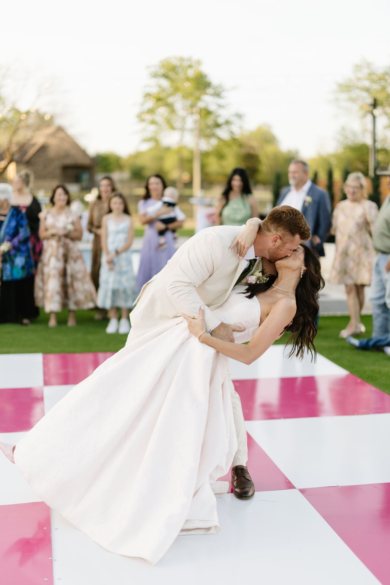 Bride and groom share a romantic dip kiss on a pink and white checkered outdoor dance floor.