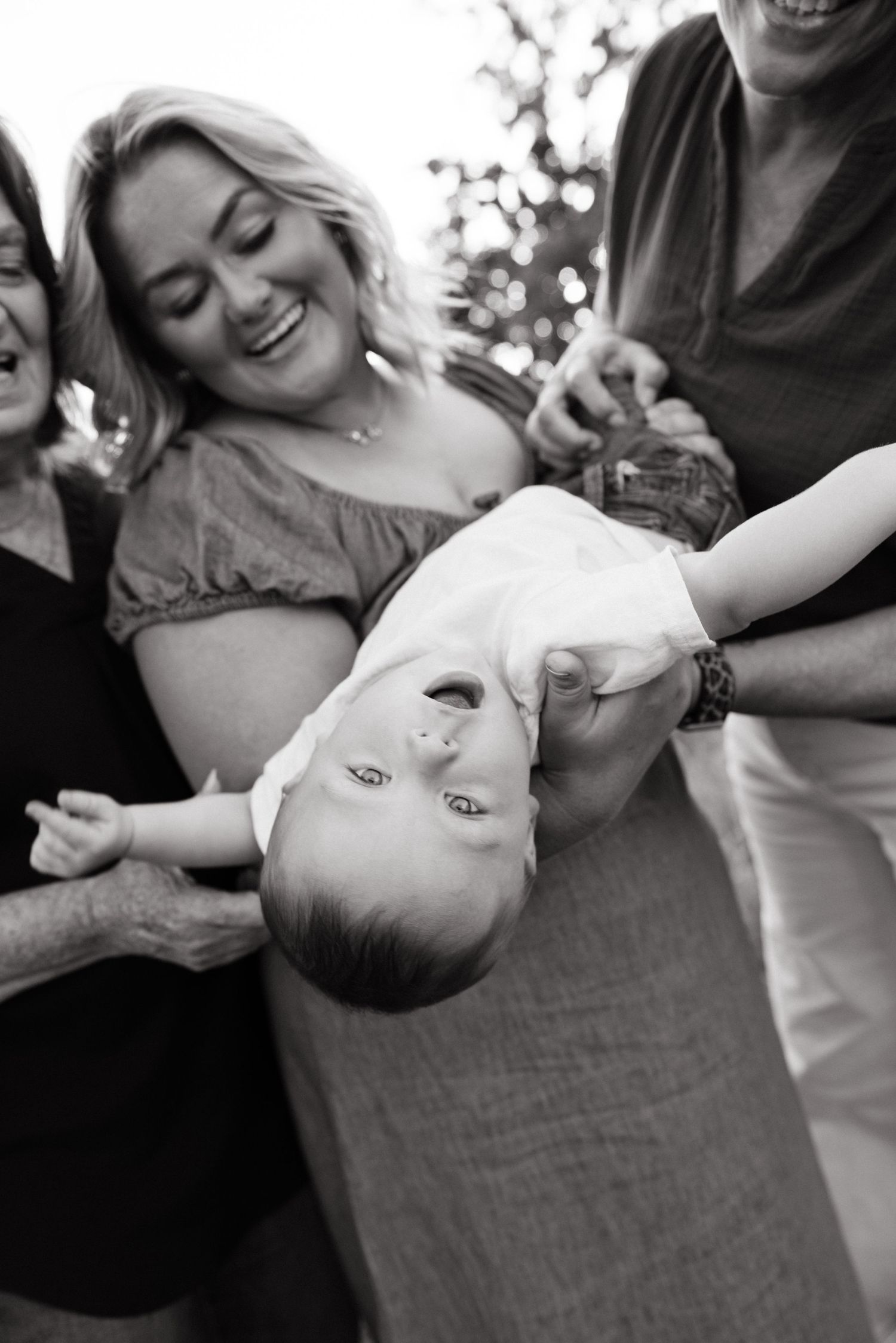 Black and white photo of a laughing family holding a smiling baby upside down outdoors.