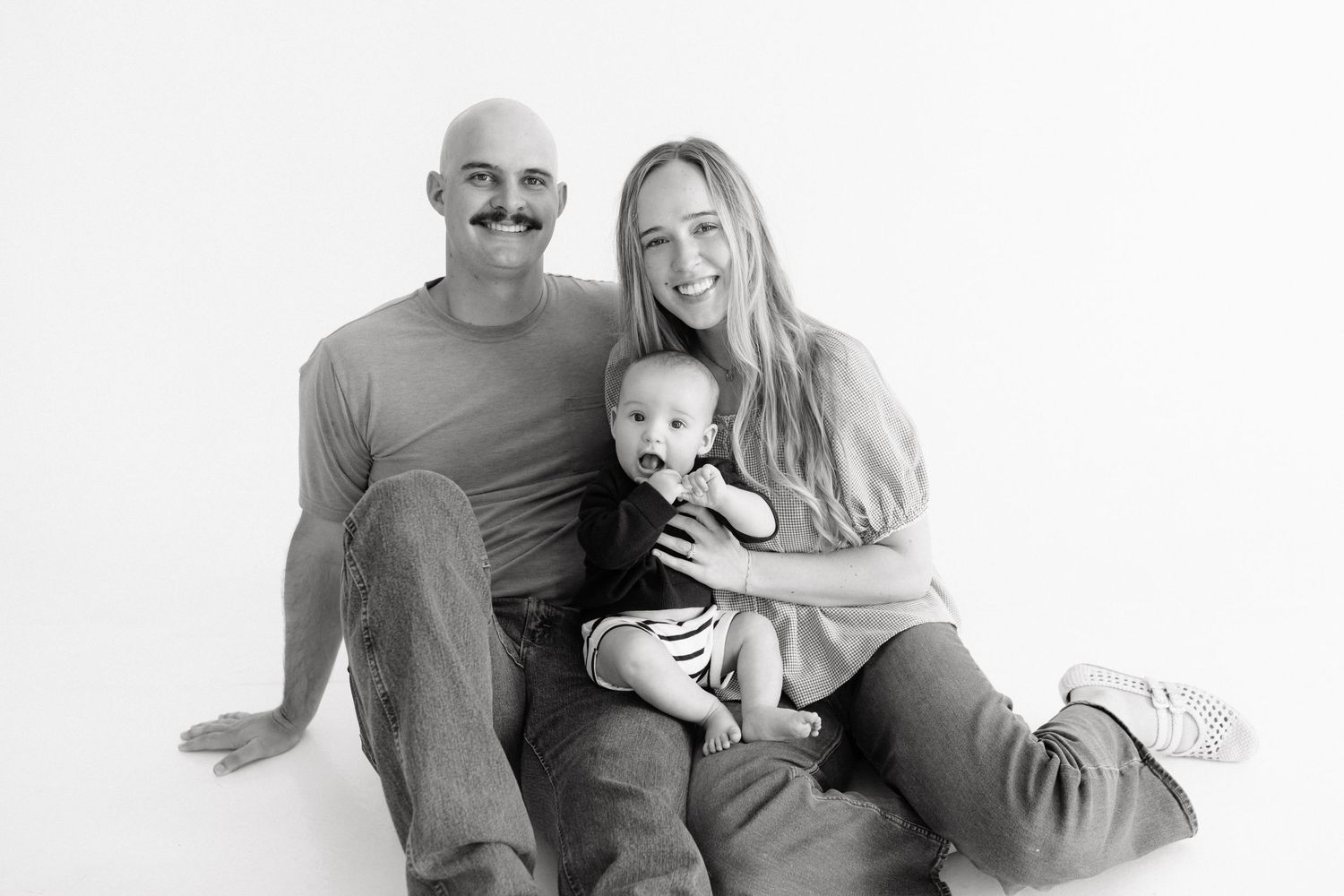 Black and white family portrait of smiling parents sitting on floor holding happy baby in a bright studio setting.