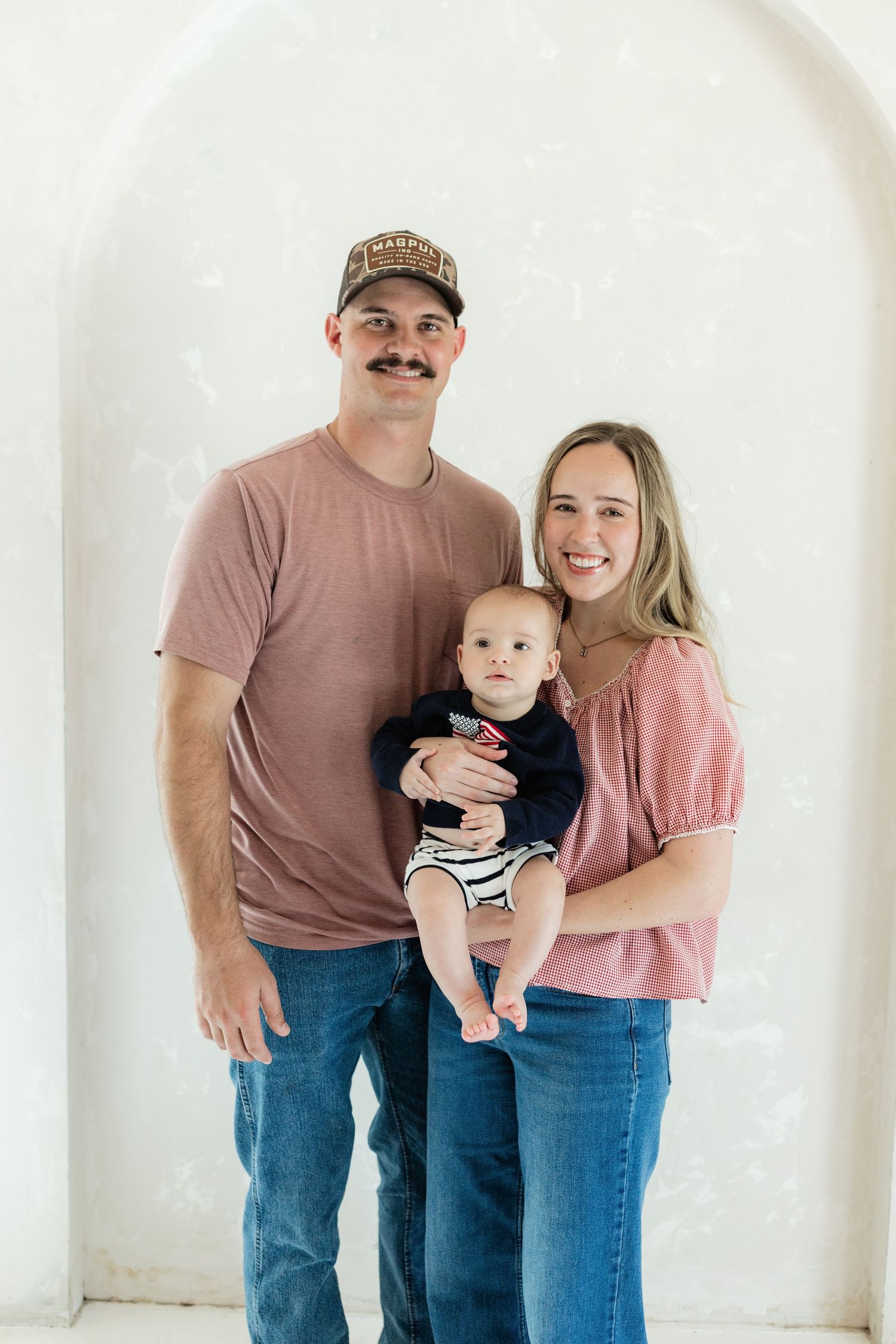 Young family portrait with father in cap, mother in pink top, and baby in navy outfit against white wall.