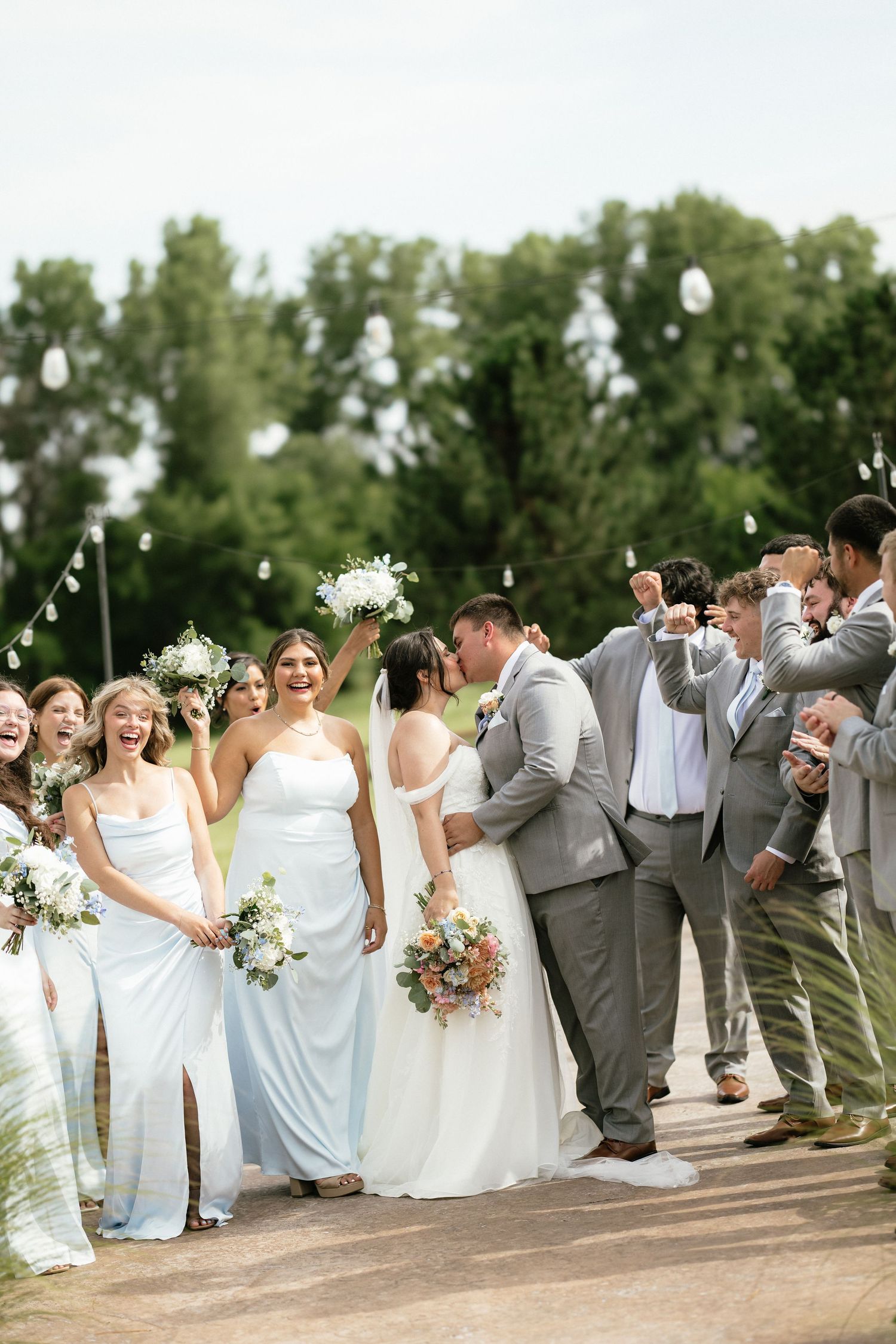 Wedding party celebrating outdoors, bride and groom kissing surrounded by bridesmaids in blue dresses and groomsmen in gray suits.