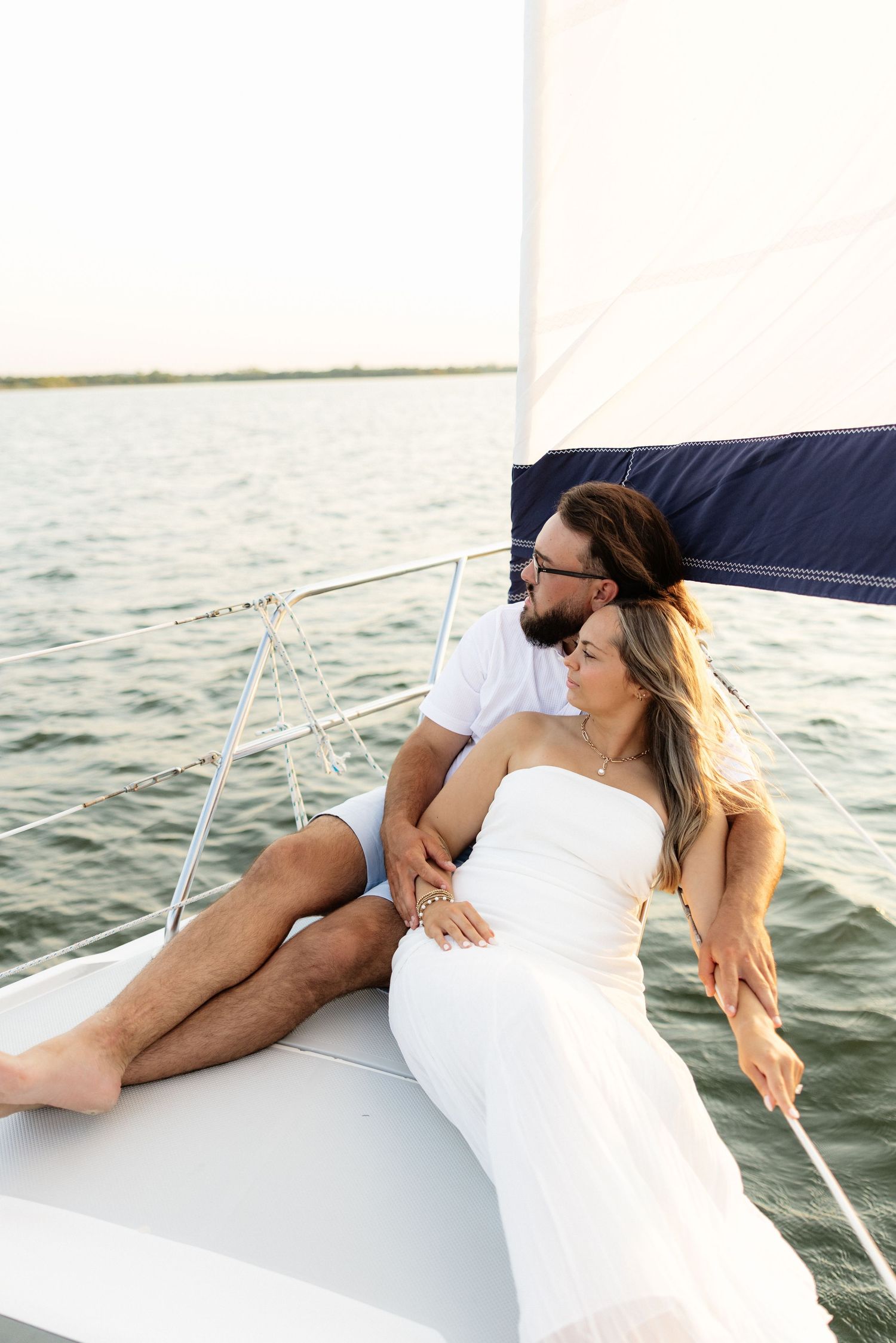 A romantic couple relaxes on a sailboat deck, woman in white dress, man in white shirt, embracing on calm water.