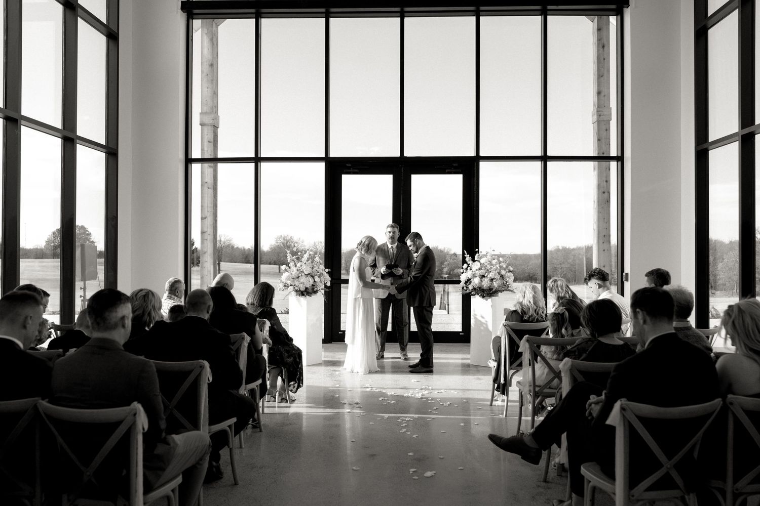 Black and white wide shot of wedding ceremony inside a bright glass-walled venue with seated guests.