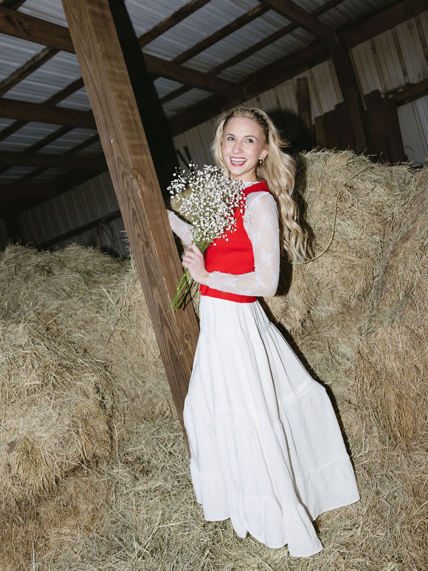 Blonde woman in red and white maxi dress holding baby's breath bouquet near hay bales in a barn.