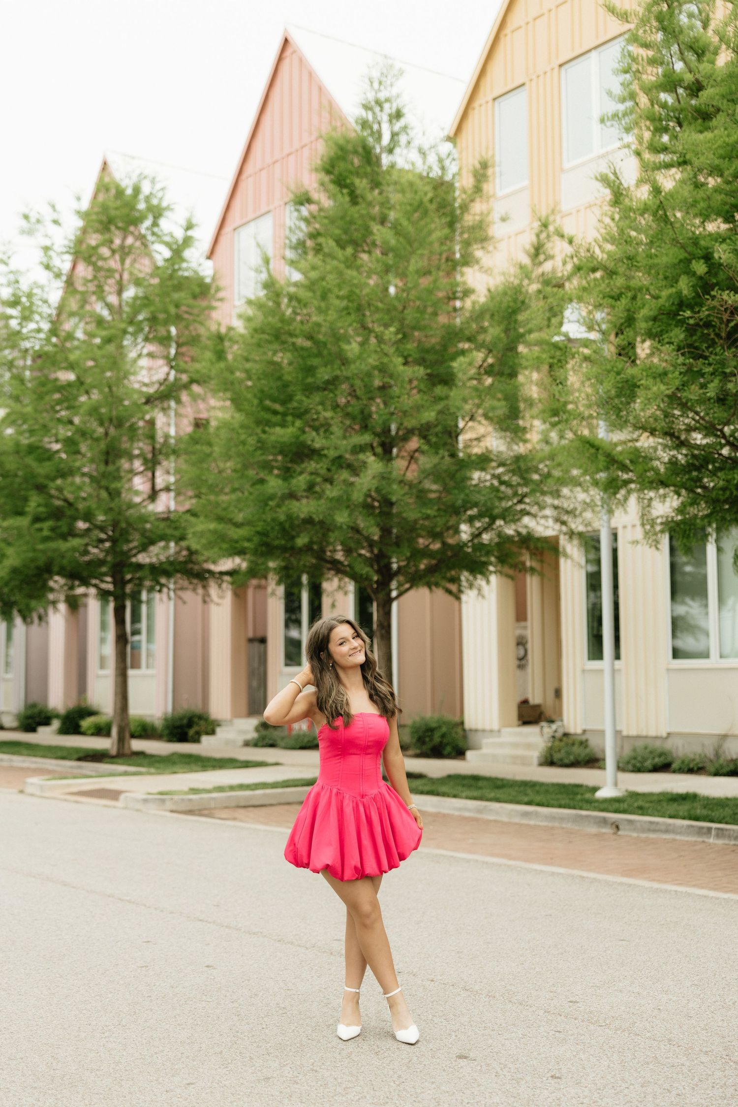 Young woman in hot pink bubble dress posing on a sunny street with green trees and modern buildings.