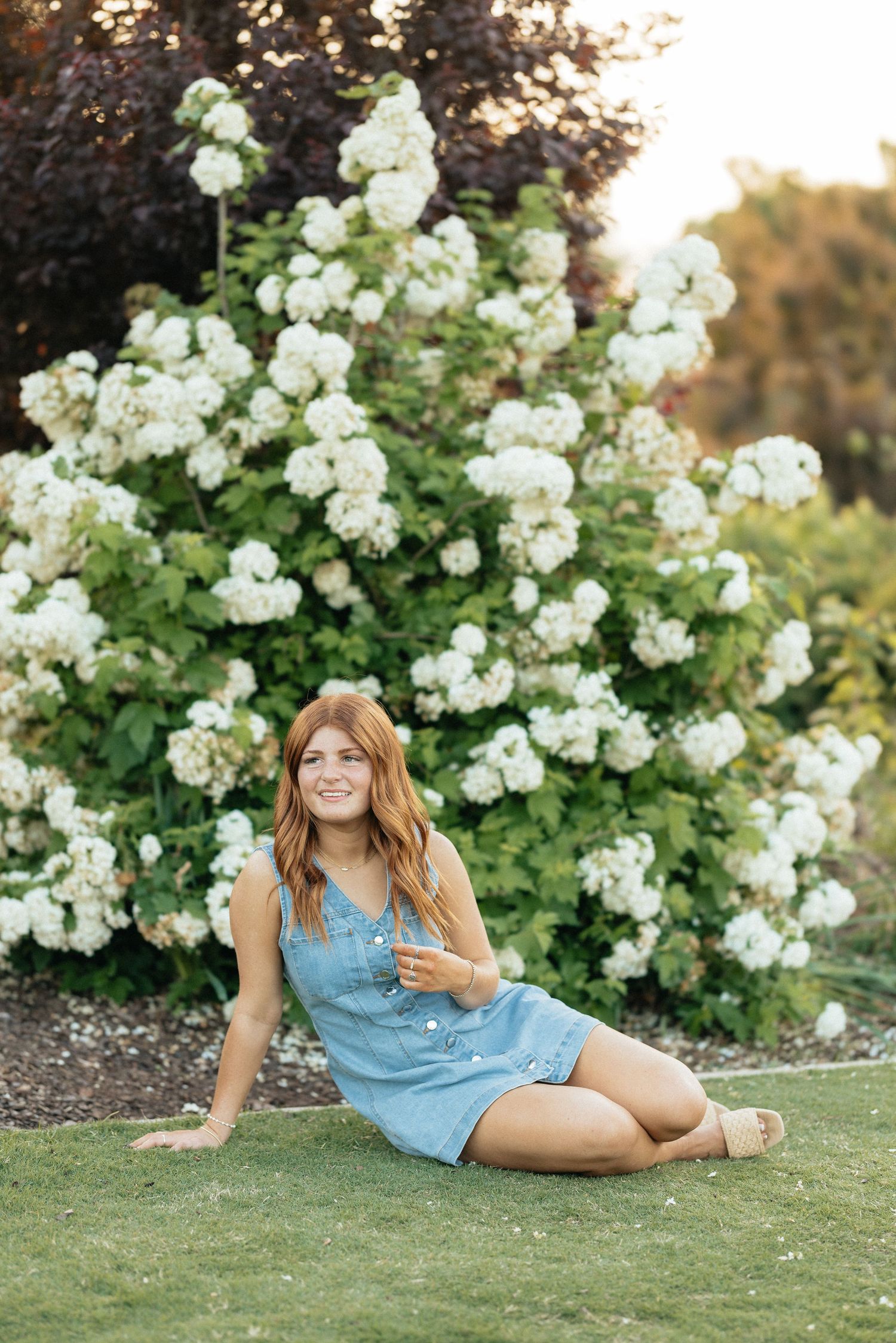 Smiling redhead teen in denim dress sits on grass beside white hydrangea bushes.