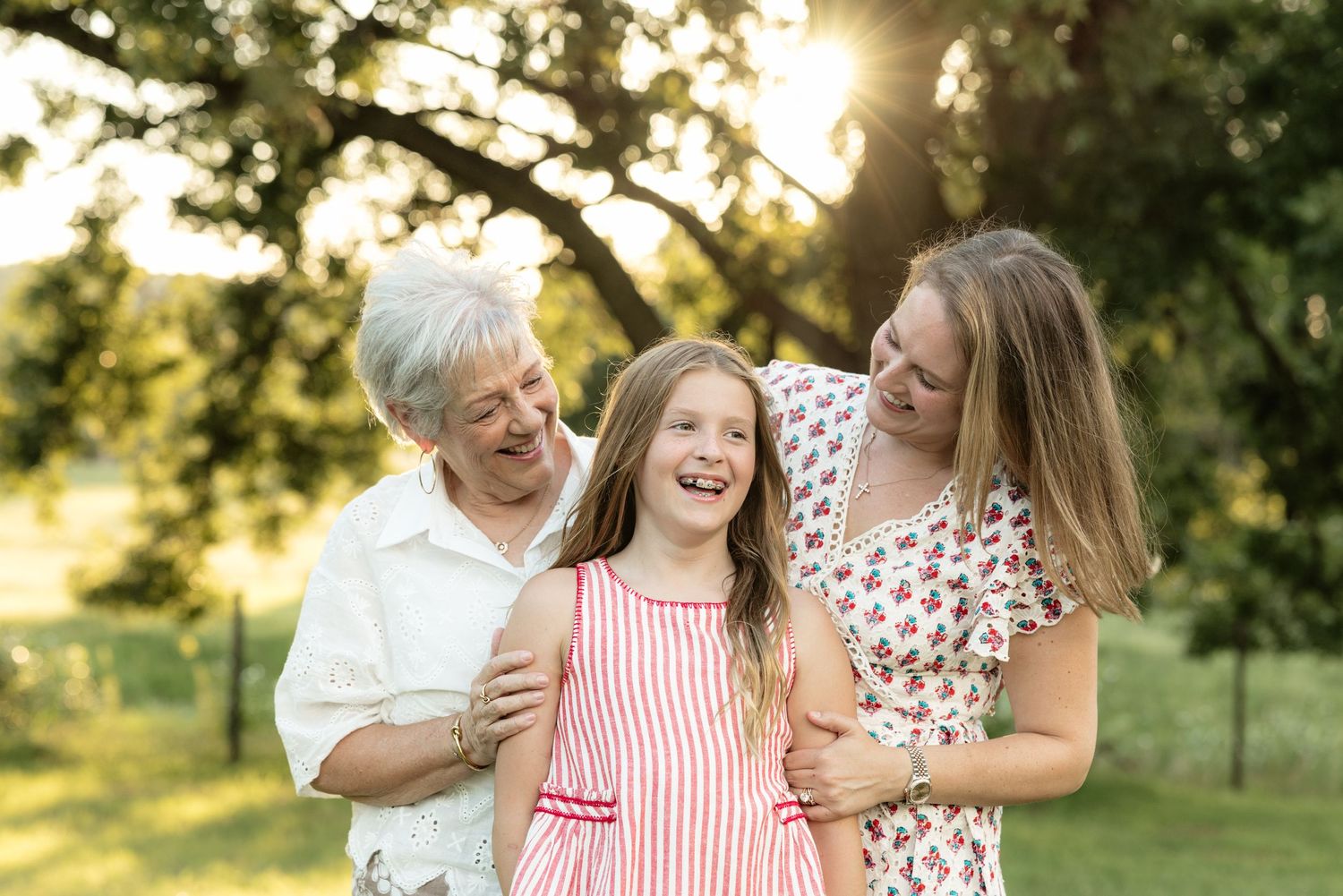 Grandmother, mother, and young girl laughing together outdoors in golden sunlight among green trees.