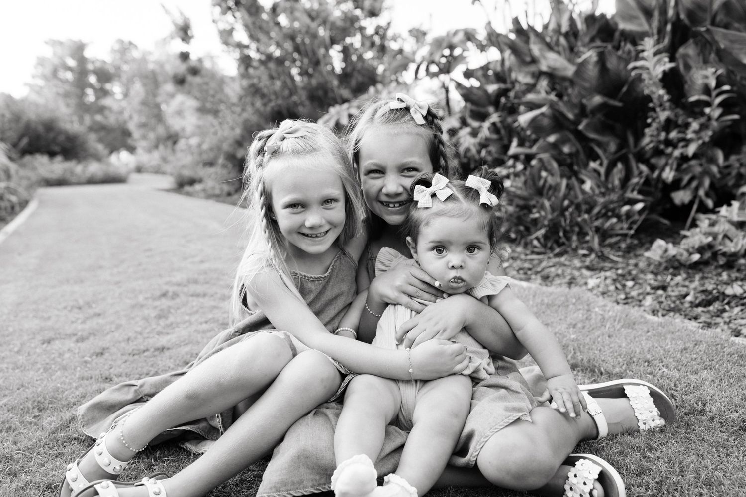 Three young girls smile together outdoors in a black and white photo, adorned with flower hair accessories.