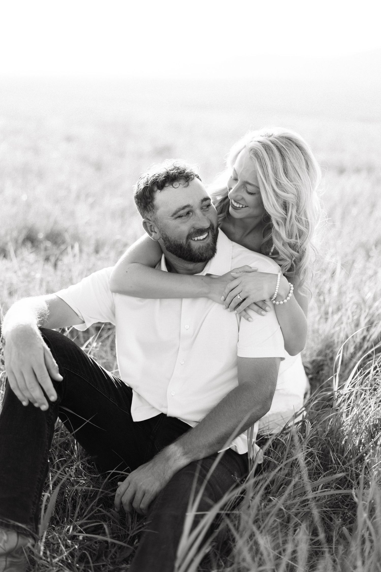 Black and white photo of a laughing couple sitting in a field, woman embracing man from behind.