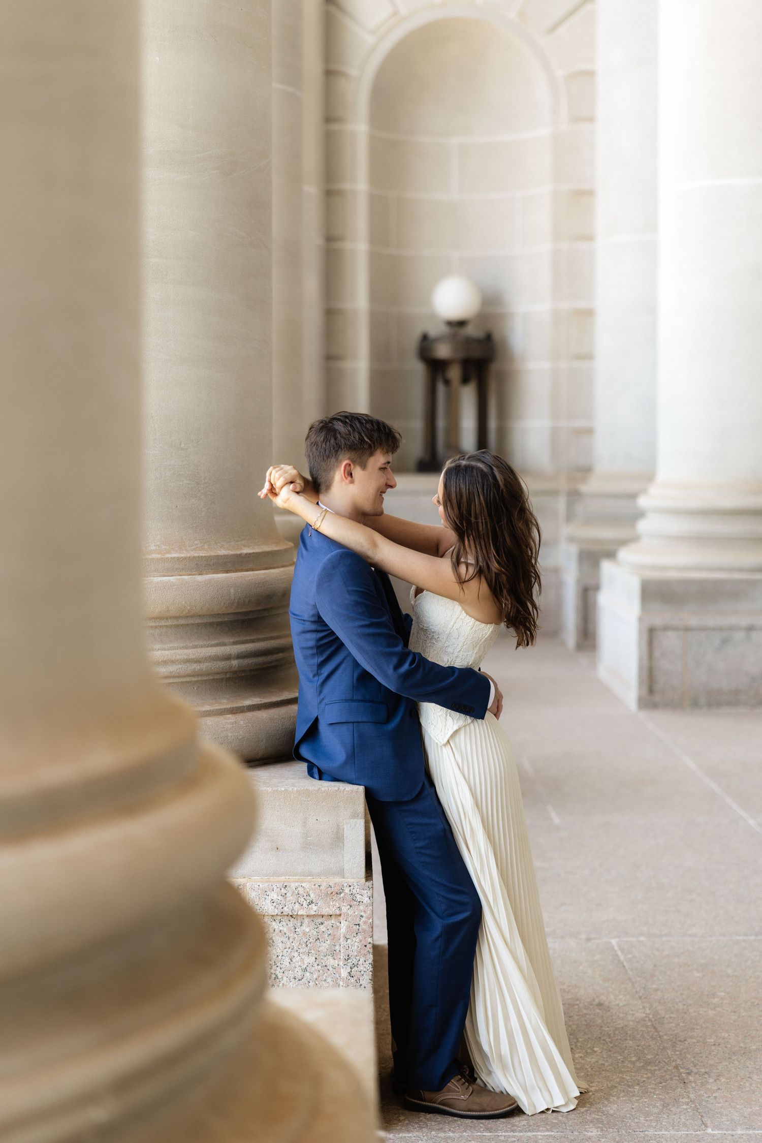 A couple embraces romantically between elegant stone columns, the groom in a blue suit and bride in a white gown.