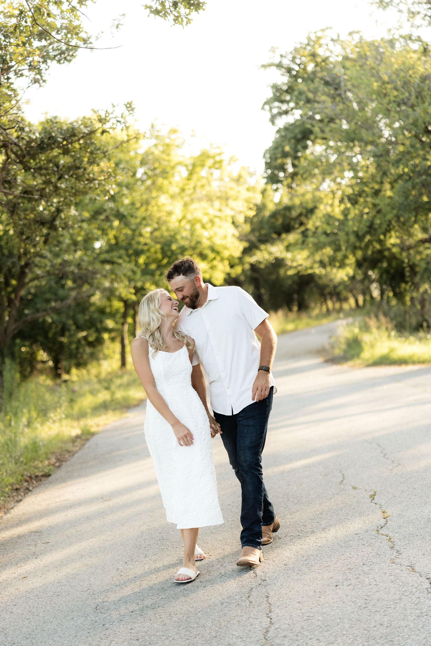 Couple walking and kissing on a sunlit road surrounded by lush green trees, woman in white dress, man in jeans.