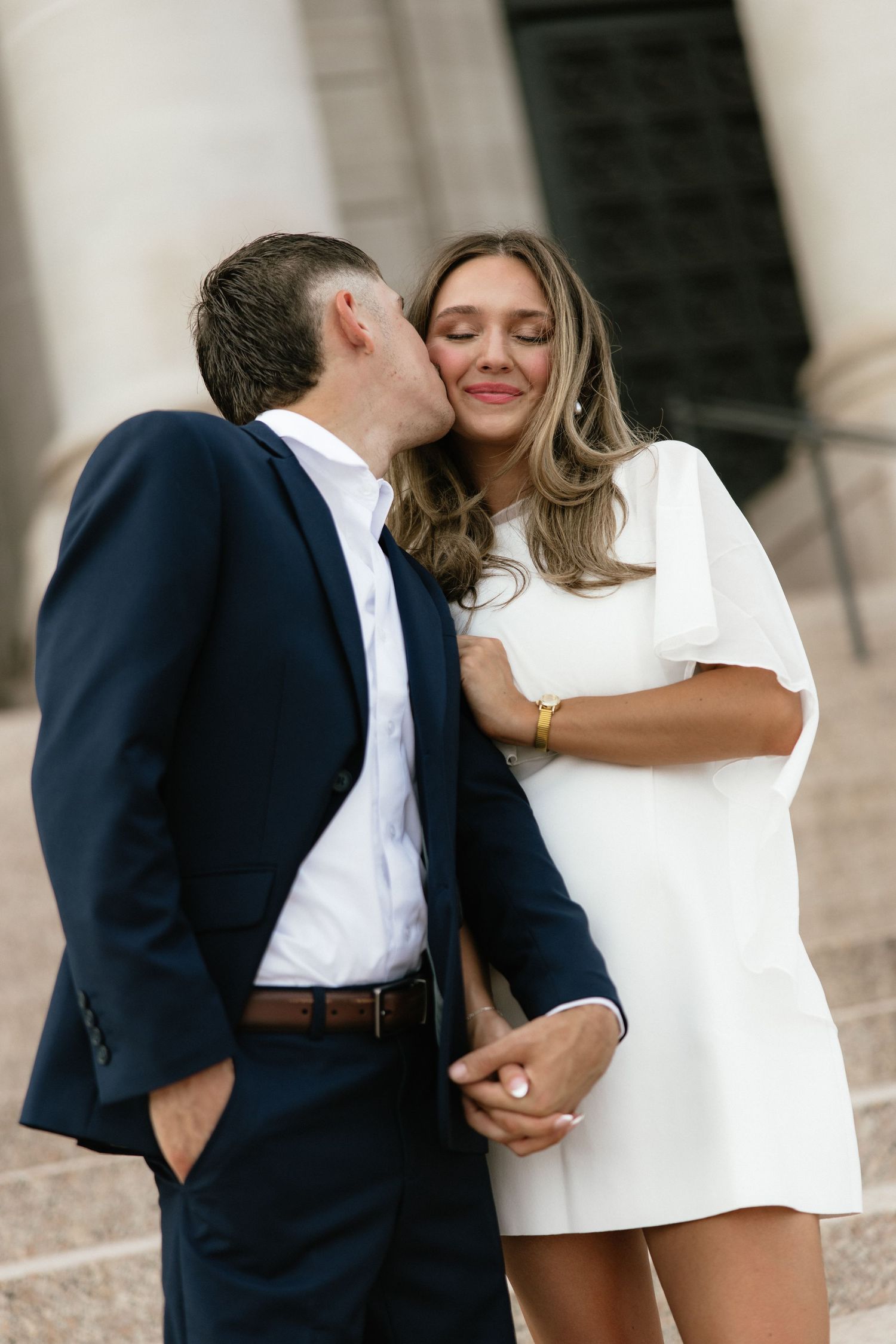 Couple sharing a kiss on steps, man in navy suit and woman in white dress.