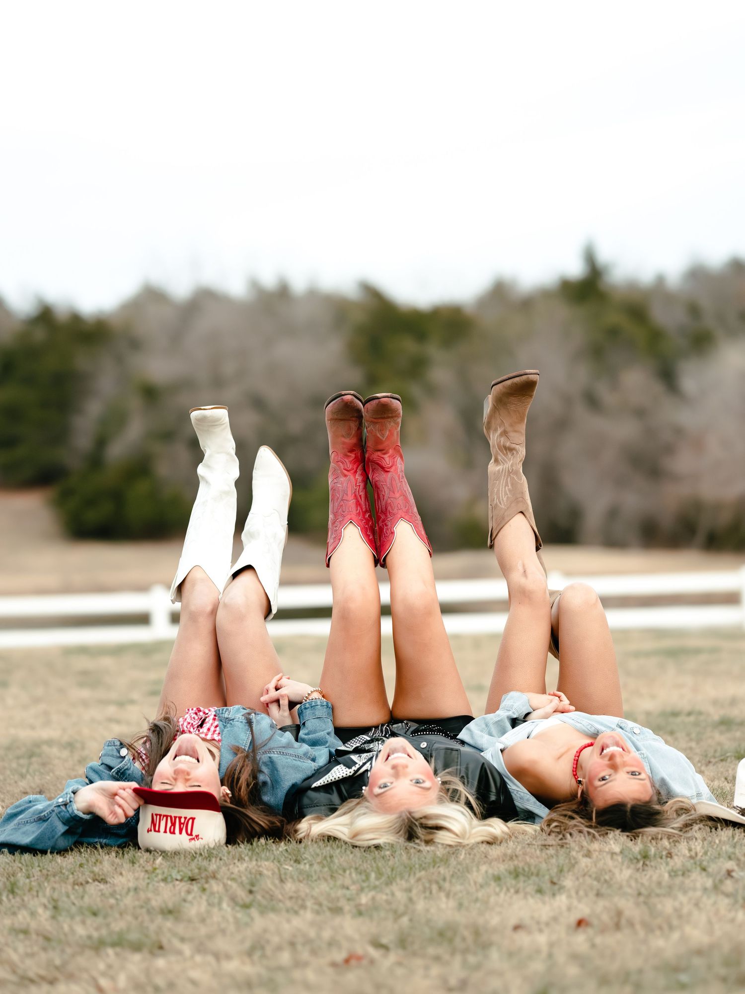 Three women lying on grass with cowboy boots raised in the air near a white fence outdoors.