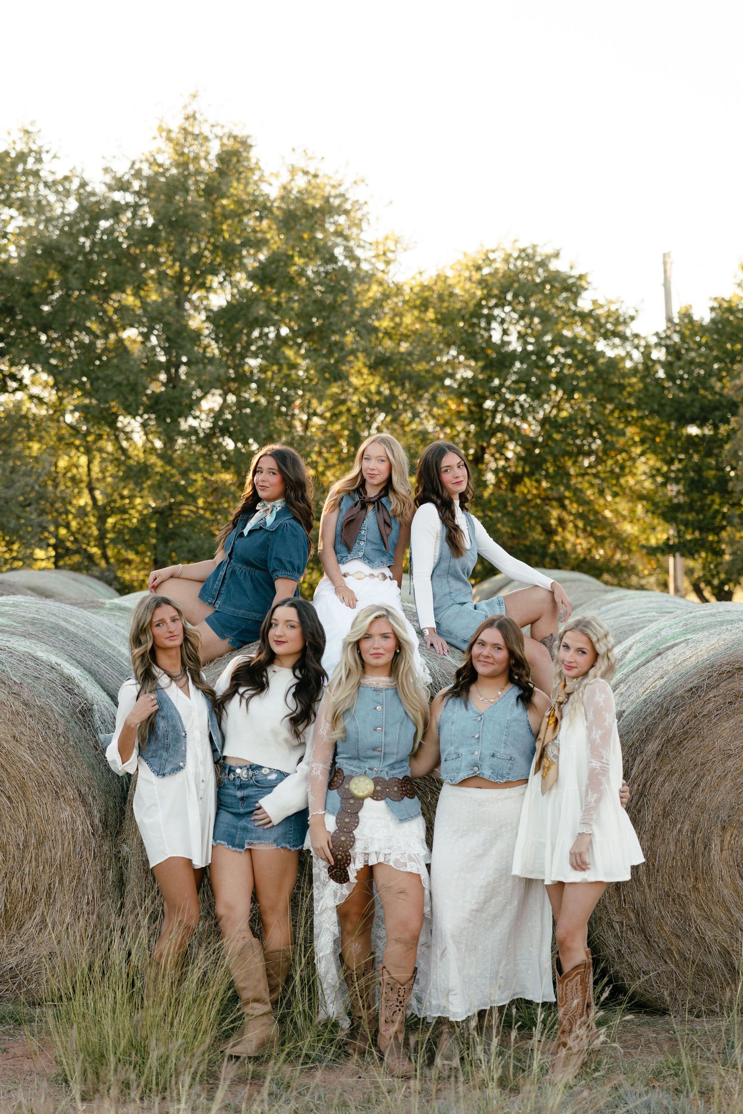 Group of seven young women in western boho outfits posing on hay bales at sunset.