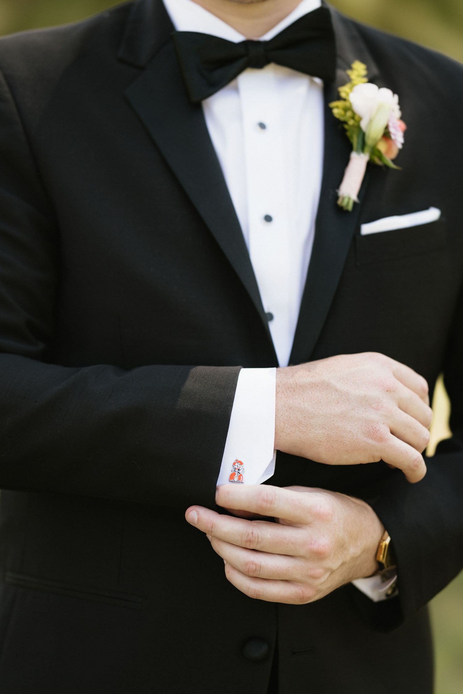 Groom in black tuxedo adjusting cufflinks with boutonniere and bow tie on wedding day.