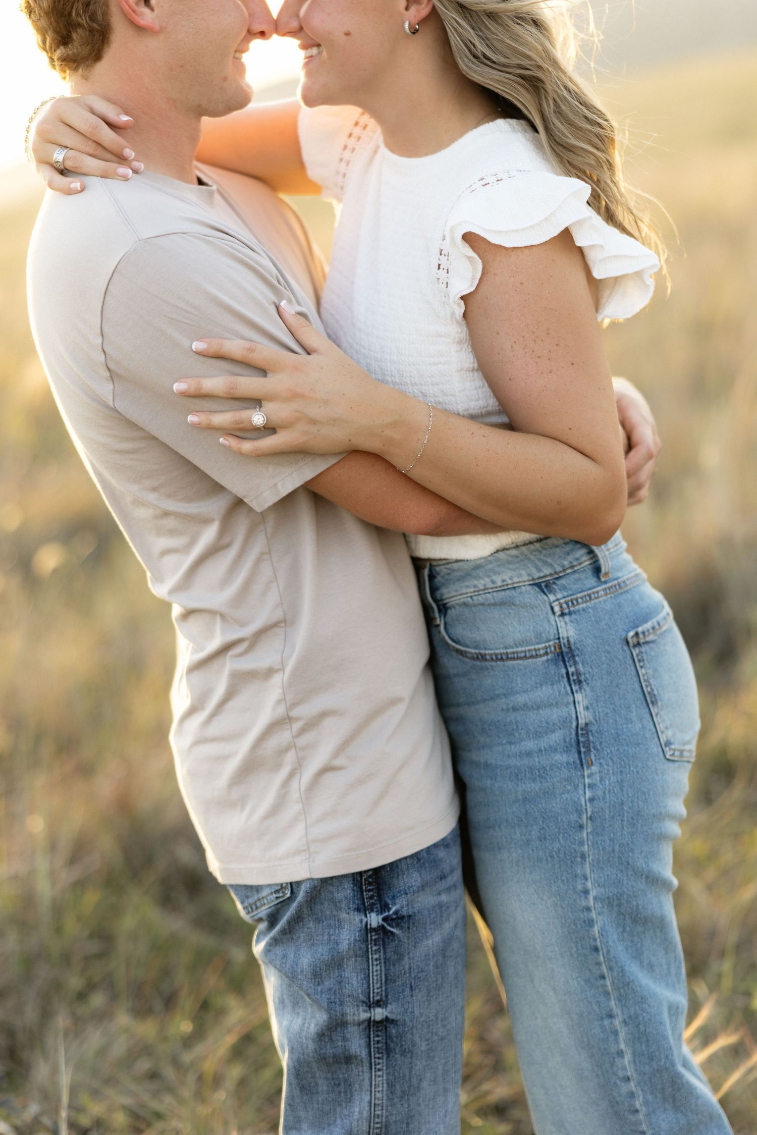 Couple embracing and almost kissing in a golden field during sunset, wearing casual jeans and light tops.