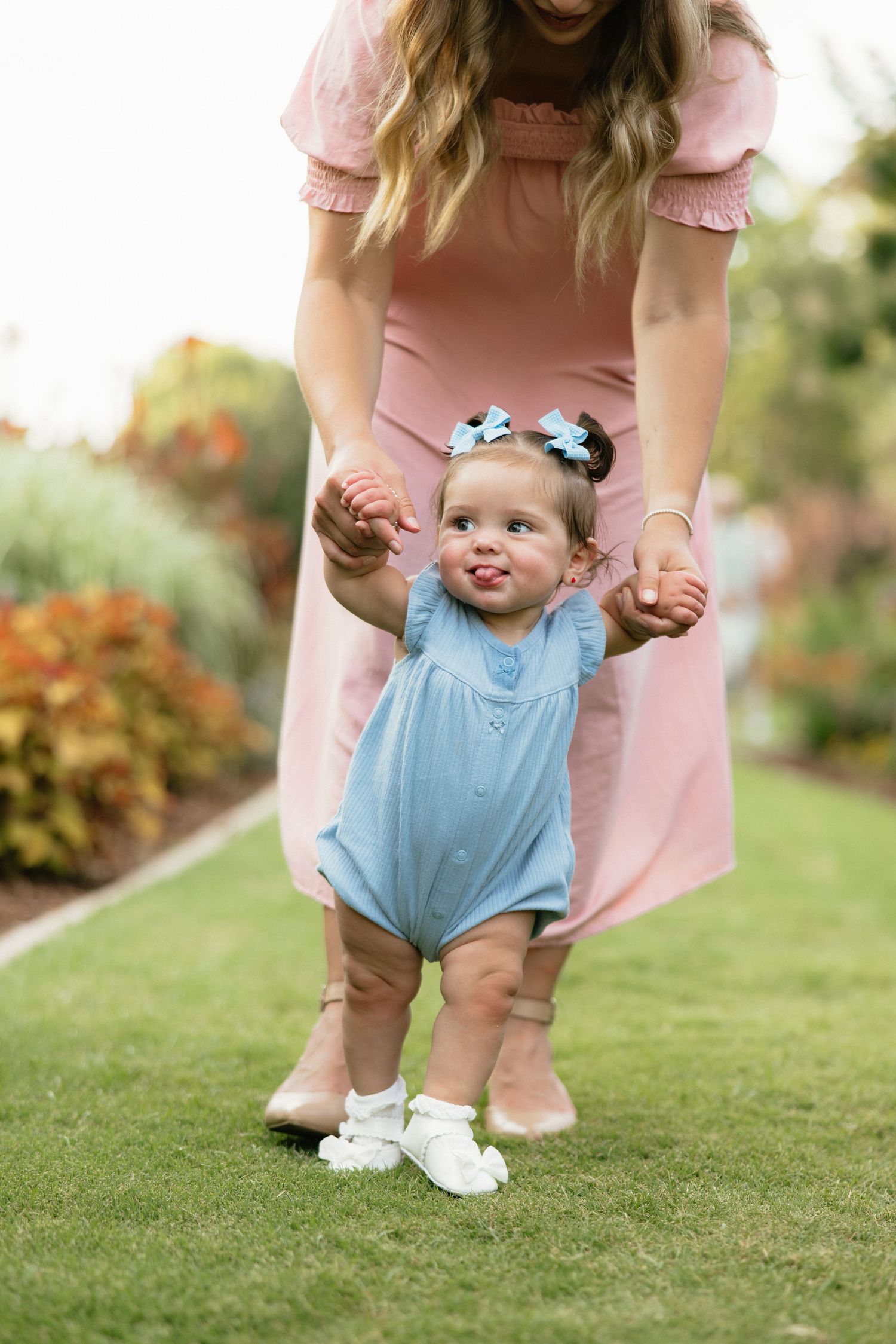 Mother in pink dress helping toddler girl in blue romper take first steps on green grass in garden.