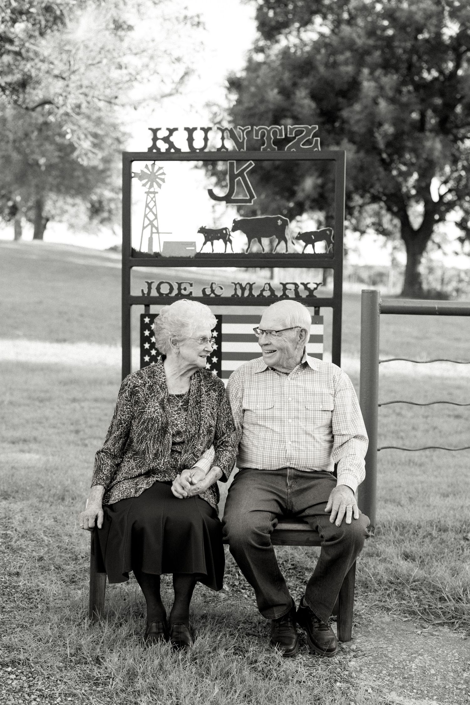 Elderly couple Joe and Mary Kuntz sit together on a bench, holding hands and smiling at each other outdoors.