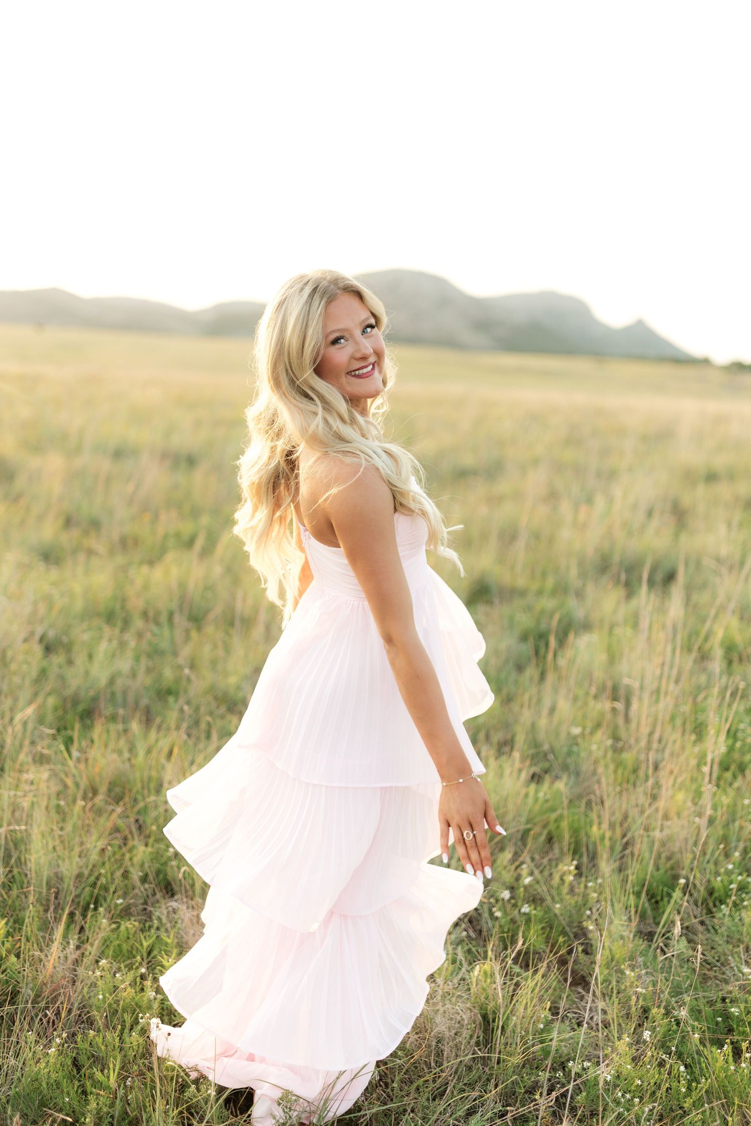 Smiling blonde woman in a flowing pink tiered dress standing in a sunlit green field with mountains in the background.