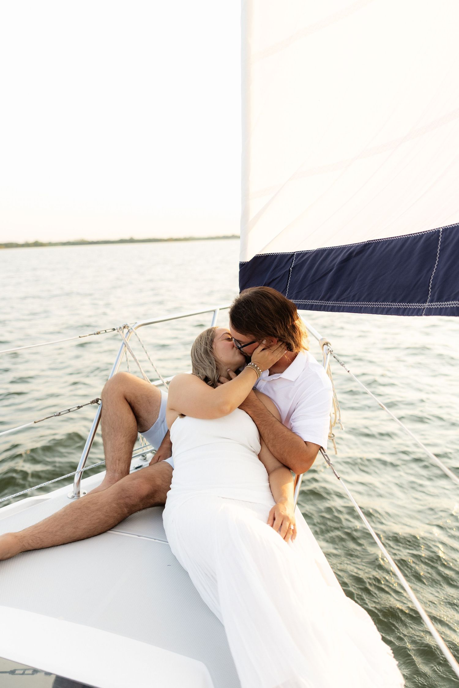 A romantic couple kissing on the bow of a sailboat on calm water, woman in white dress, man in white shirt.