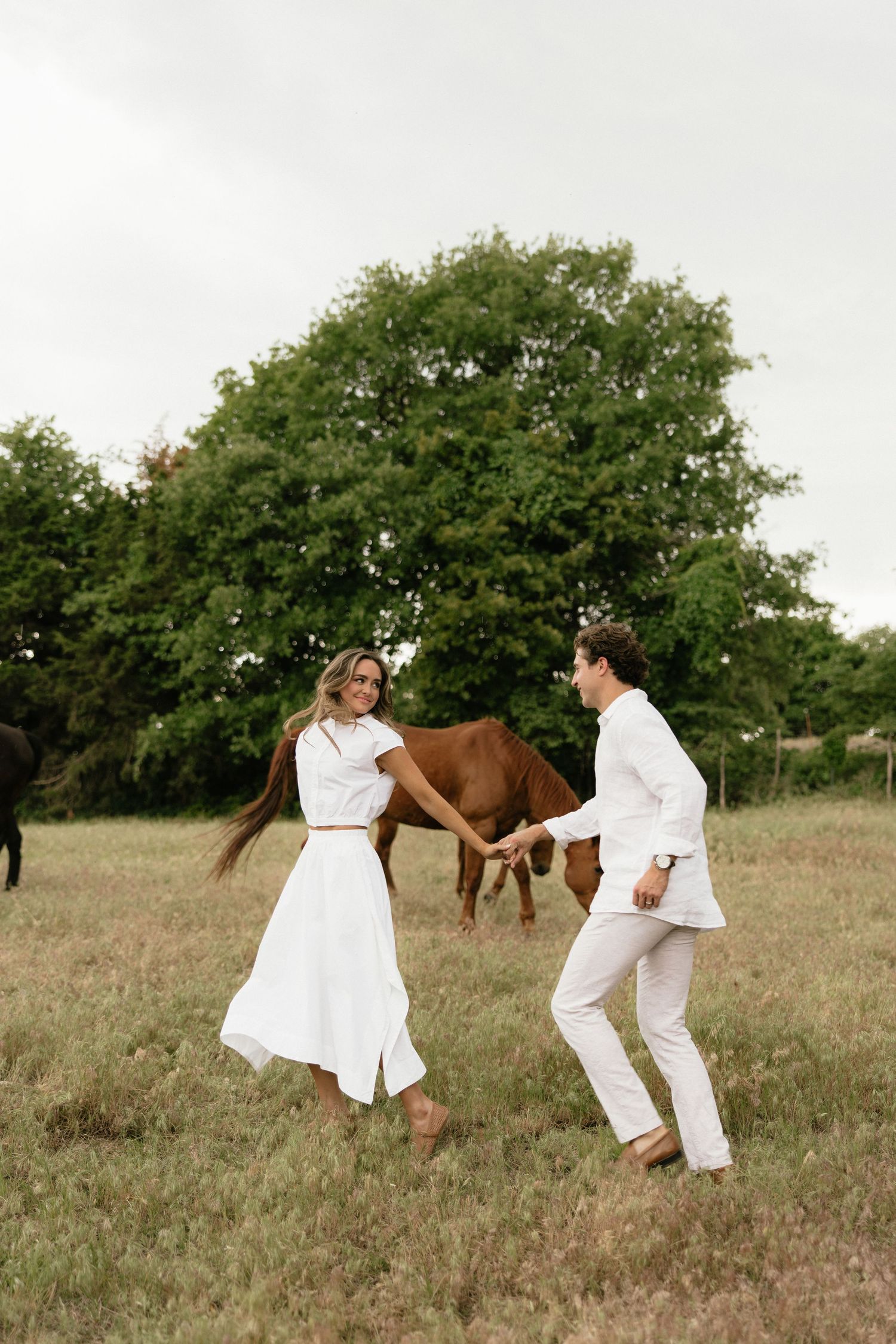 Couple in white holding hands dancing in green field with horse and trees in background.