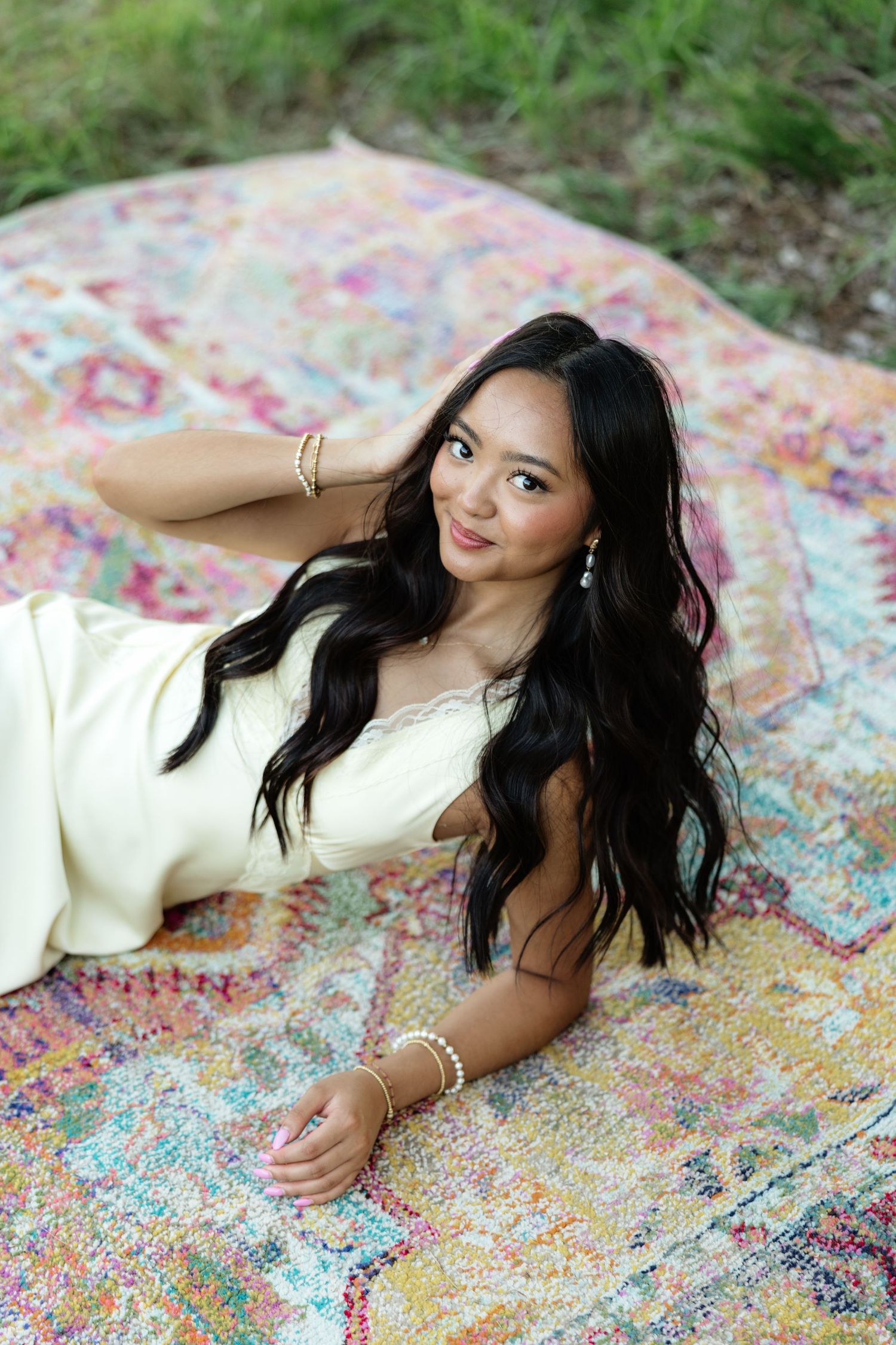 Young woman in yellow dress lying on colorful boho rug outdoors, long black hair, gold jewelry, smiling.