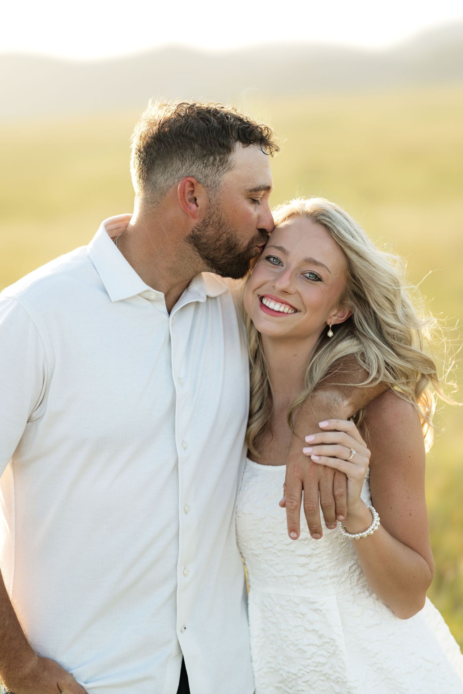 A couple in white attire shares a romantic kiss in a golden field, with the woman smiling and showing her engagement ring.