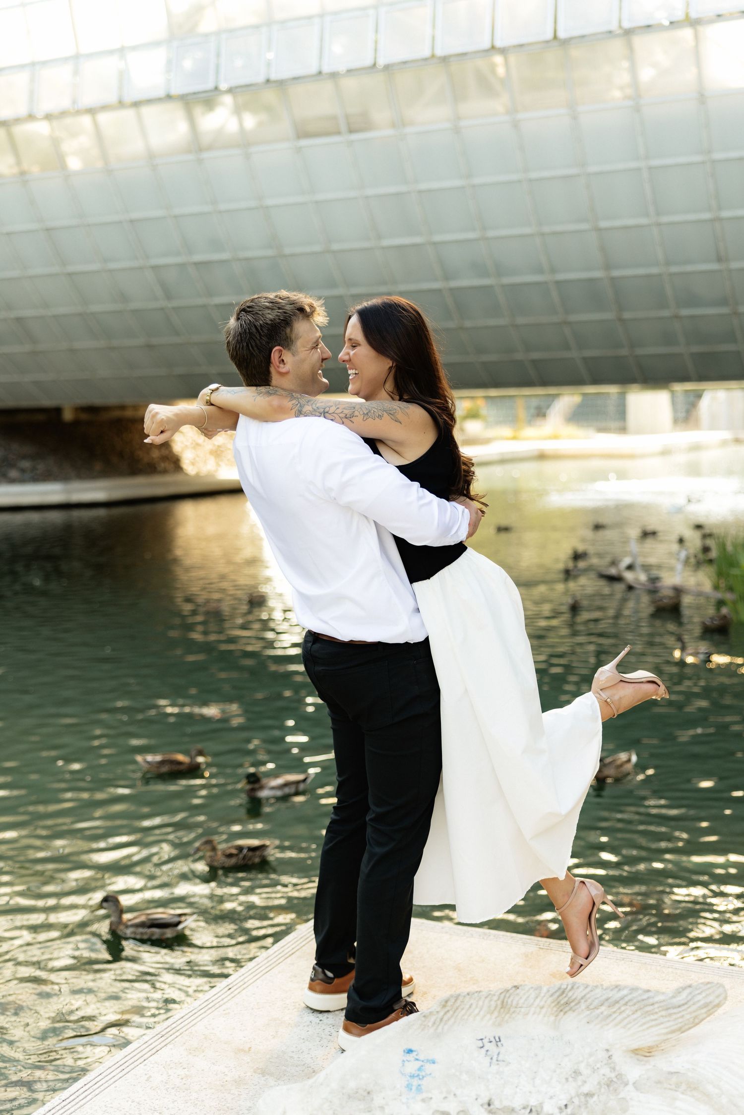 Couple embracing and dancing by a pond with ducks, woman in white skirt lifted by man in white shirt.