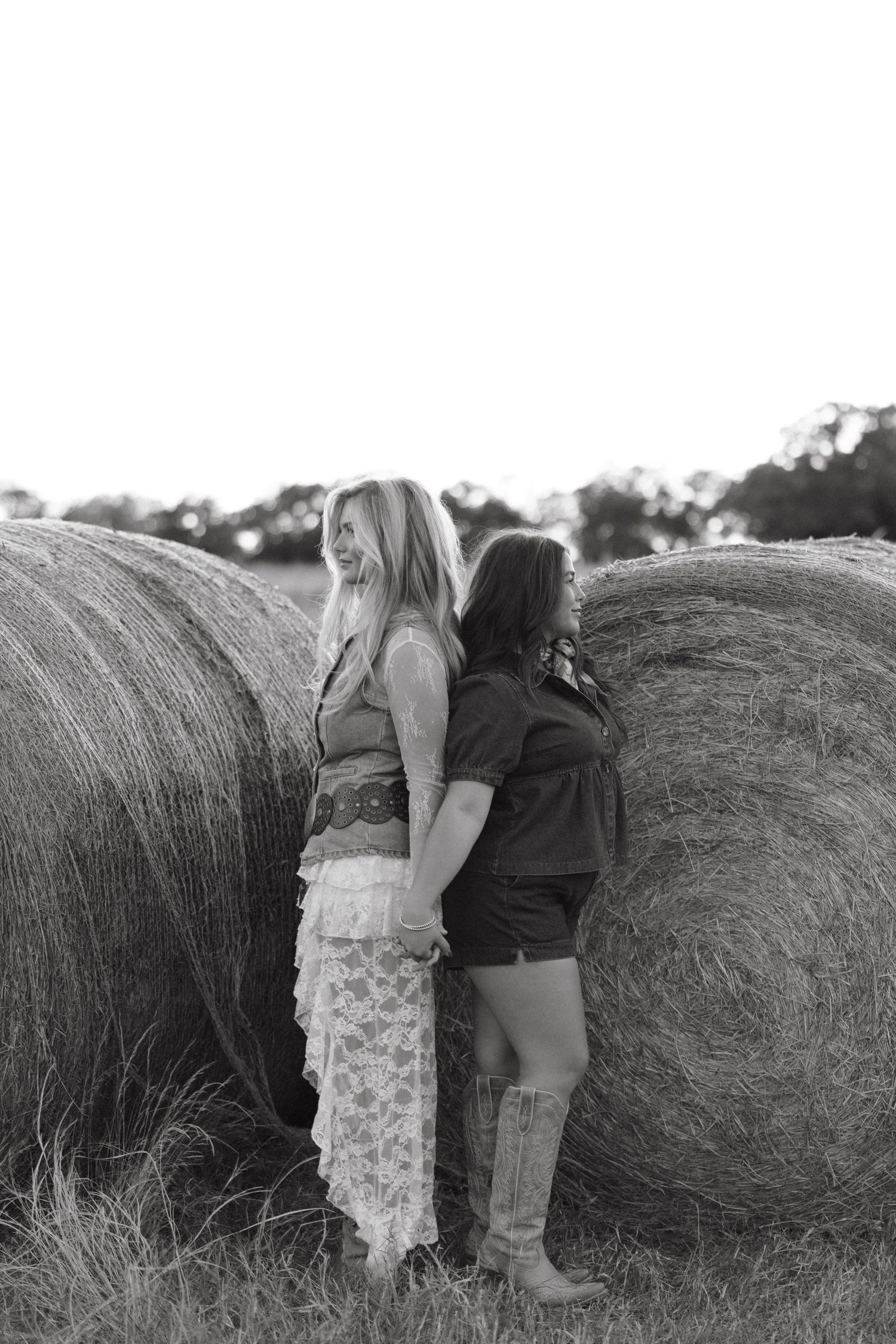 Black and white photo of two young women standing back to back against large hay bales in a field.