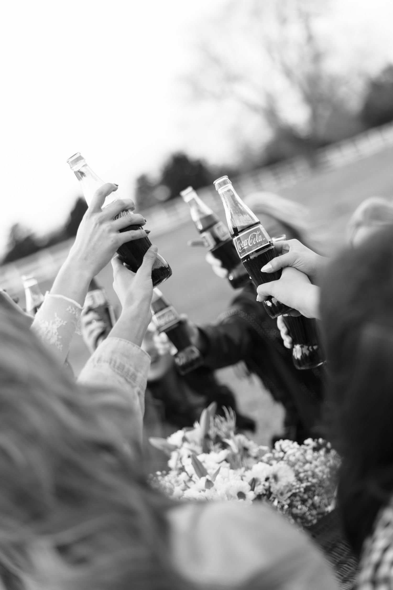 Black and white close-up of hands raising Coca-Cola bottles in a toast at outdoor celebration.
