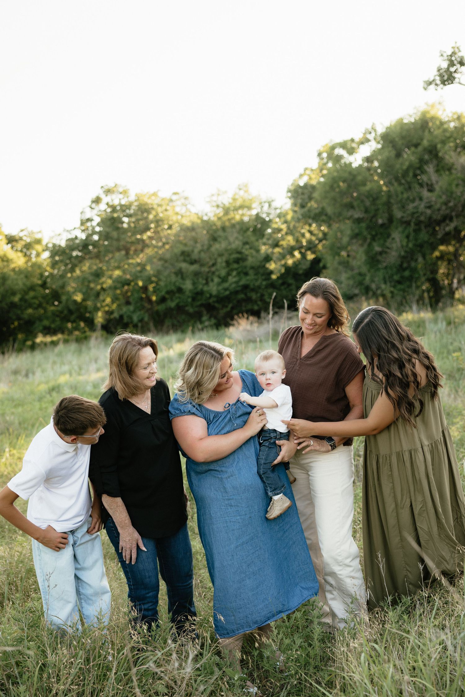 Family gathering outdoors in a green meadow, with women admiring a baby held by one member, and a young boy nearby.