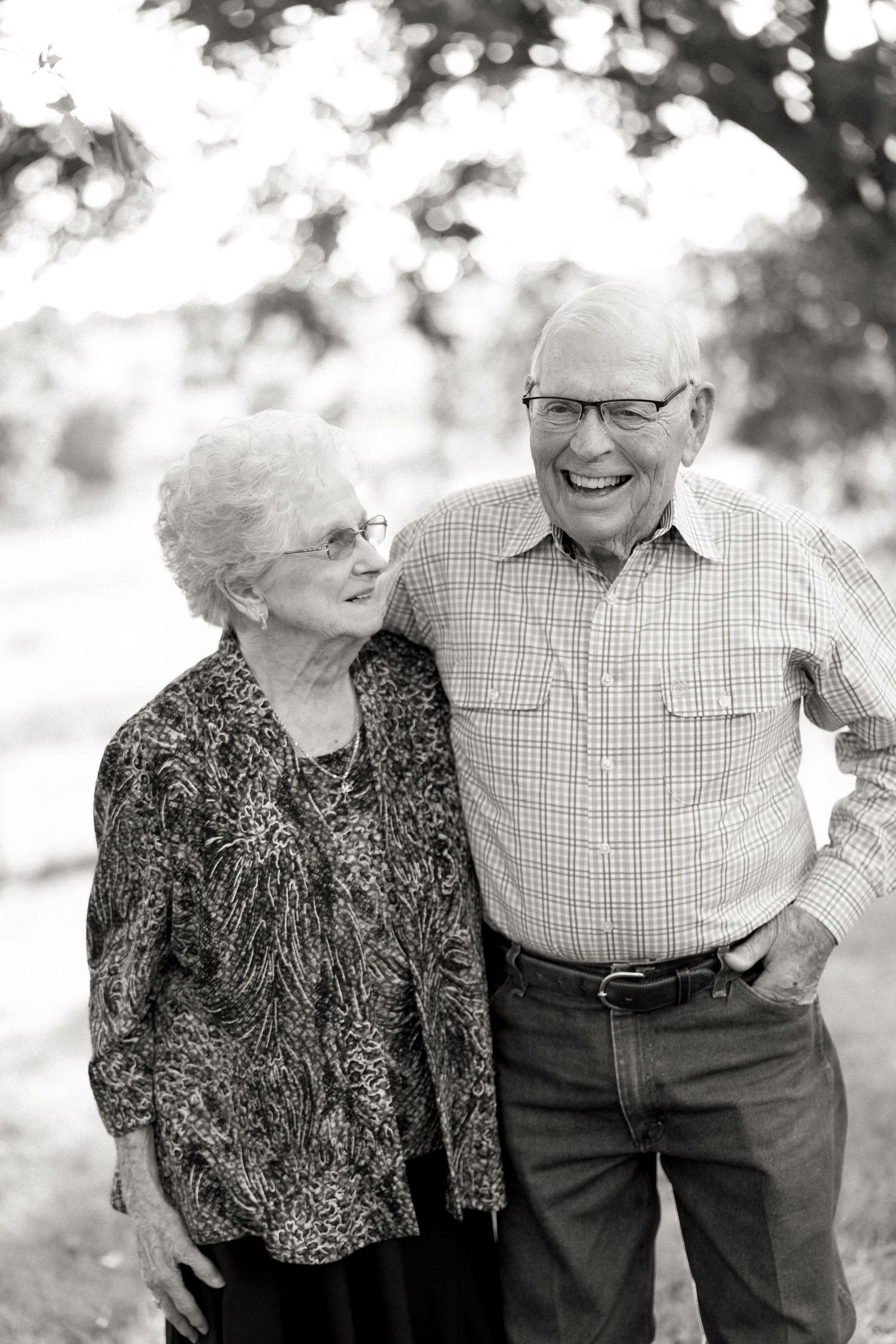 Black and white portrait of a smiling elderly couple embracing outdoors, wearing glasses and casual clothing.