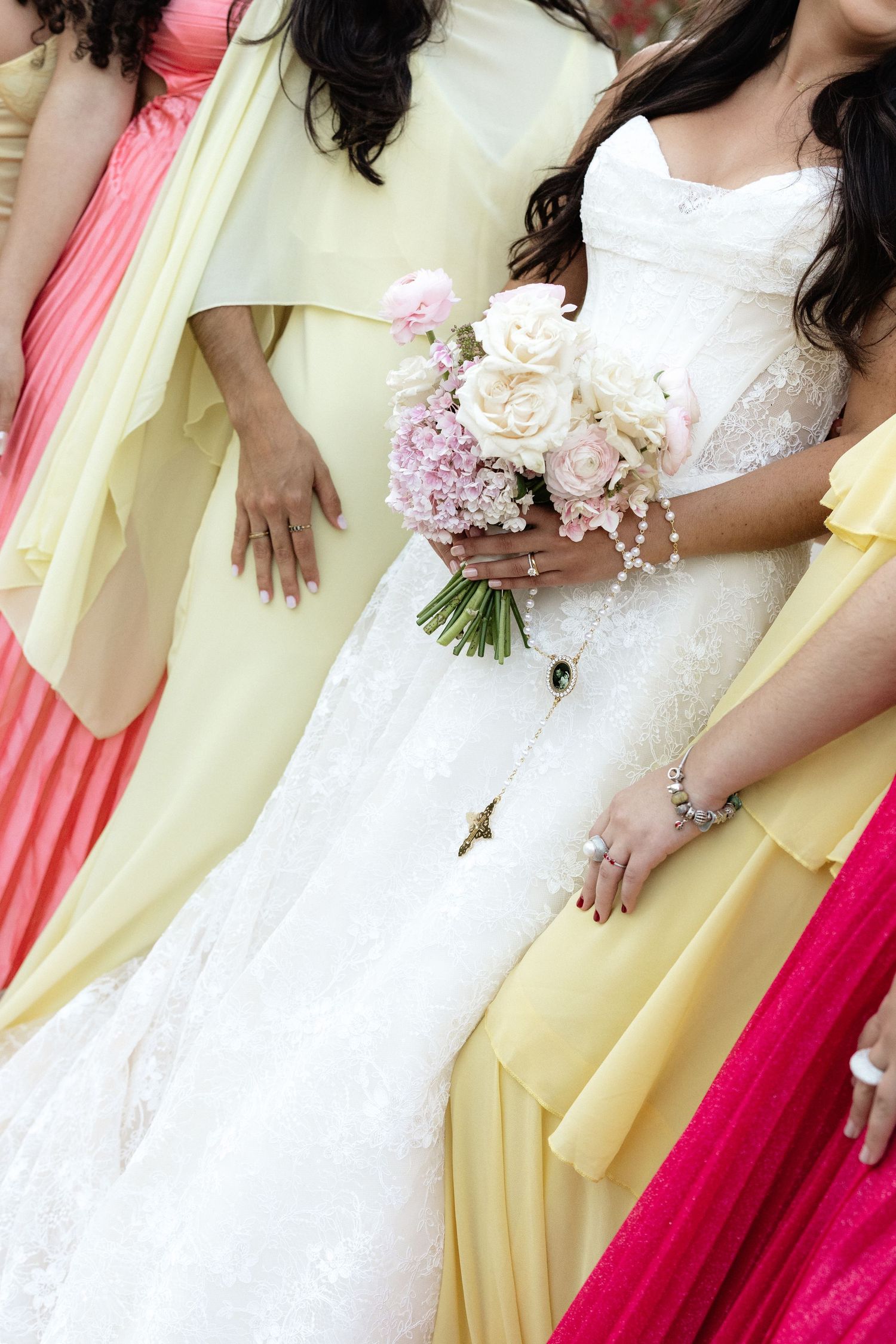 Bride in white lace gown holding pink and cream bouquet, flanked by bridesmaids in yellow and pink dresses.