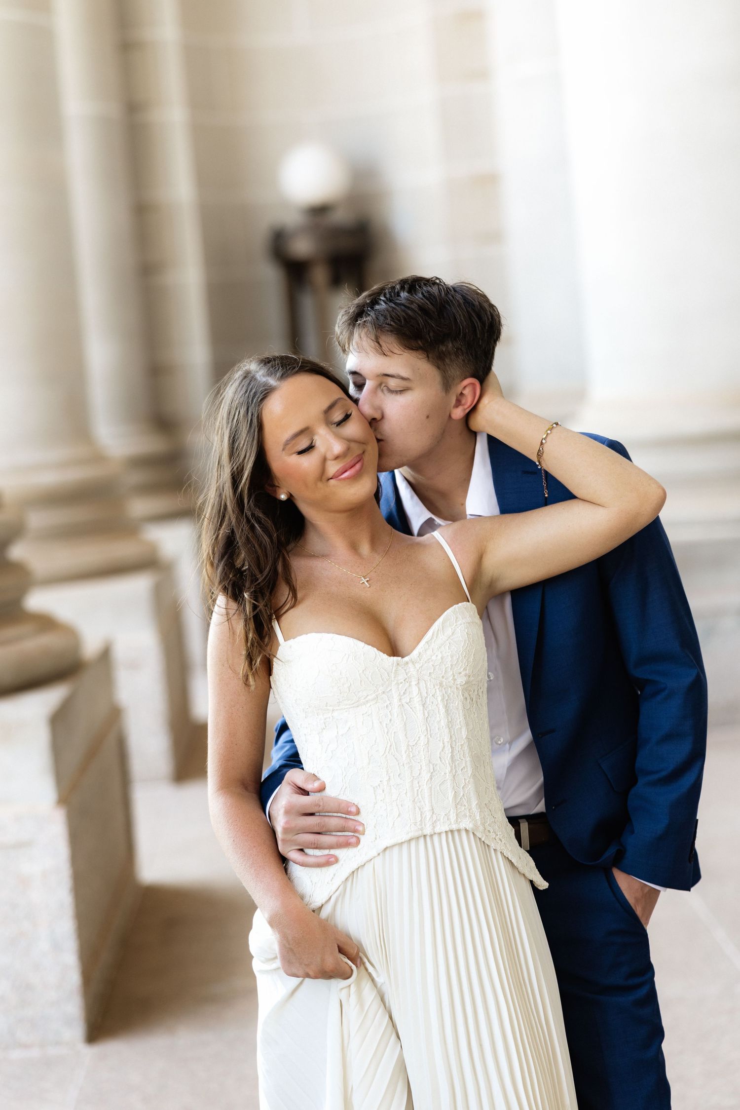 A groom in a navy suit kisses his bride on the cheek as she smiles in a white strapless gown near elegant columns.