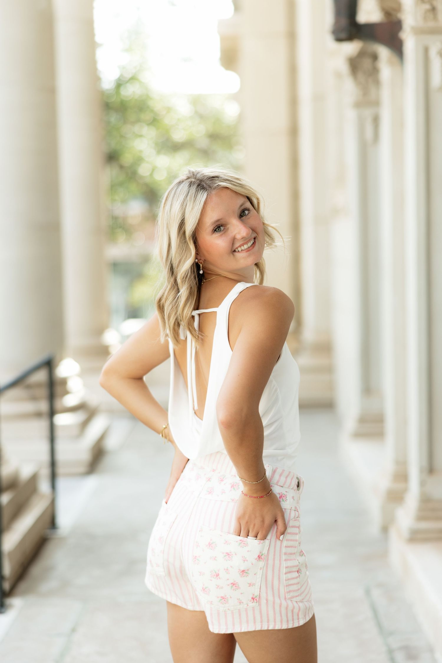 Smiling blonde woman in white tank top and floral shorts posing outdoors near elegant columns.