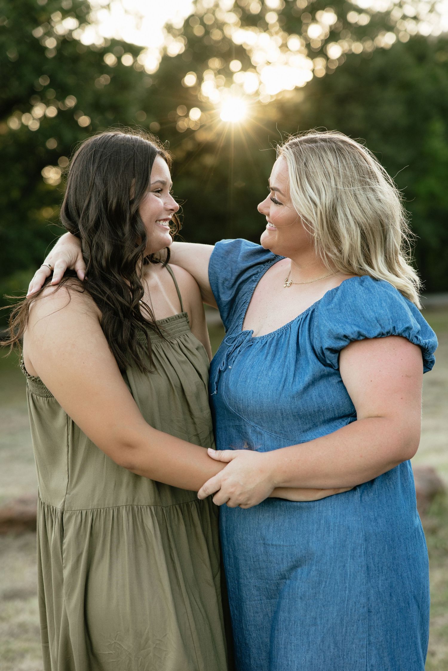 Two smiling women in summer dresses embrace outdoors at golden hour with sunlight filtering through trees.
