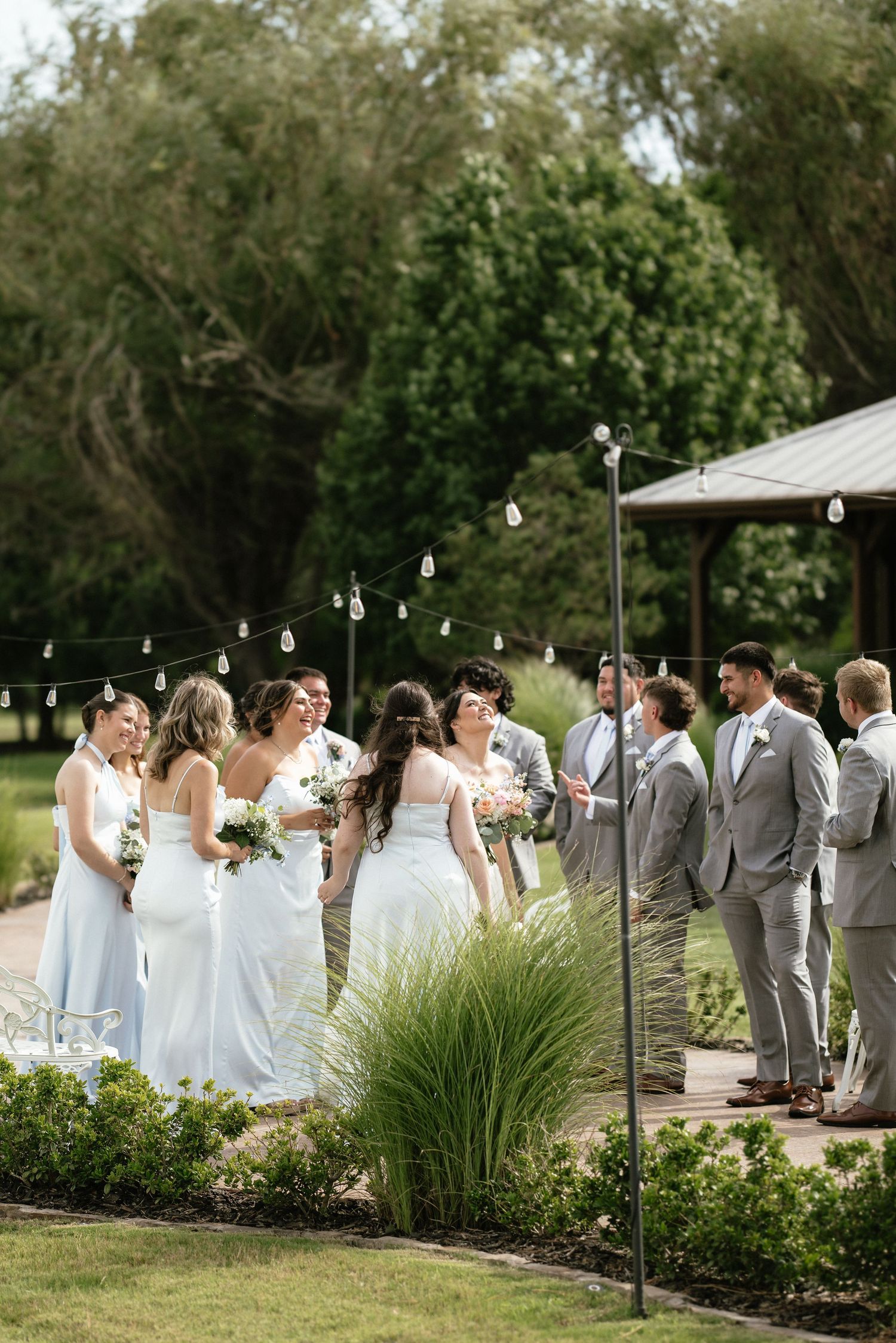 Wedding party in outdoor garden setting, bridesmaids in light blue gowns and groomsmen in gray suits, string lights overhead.
