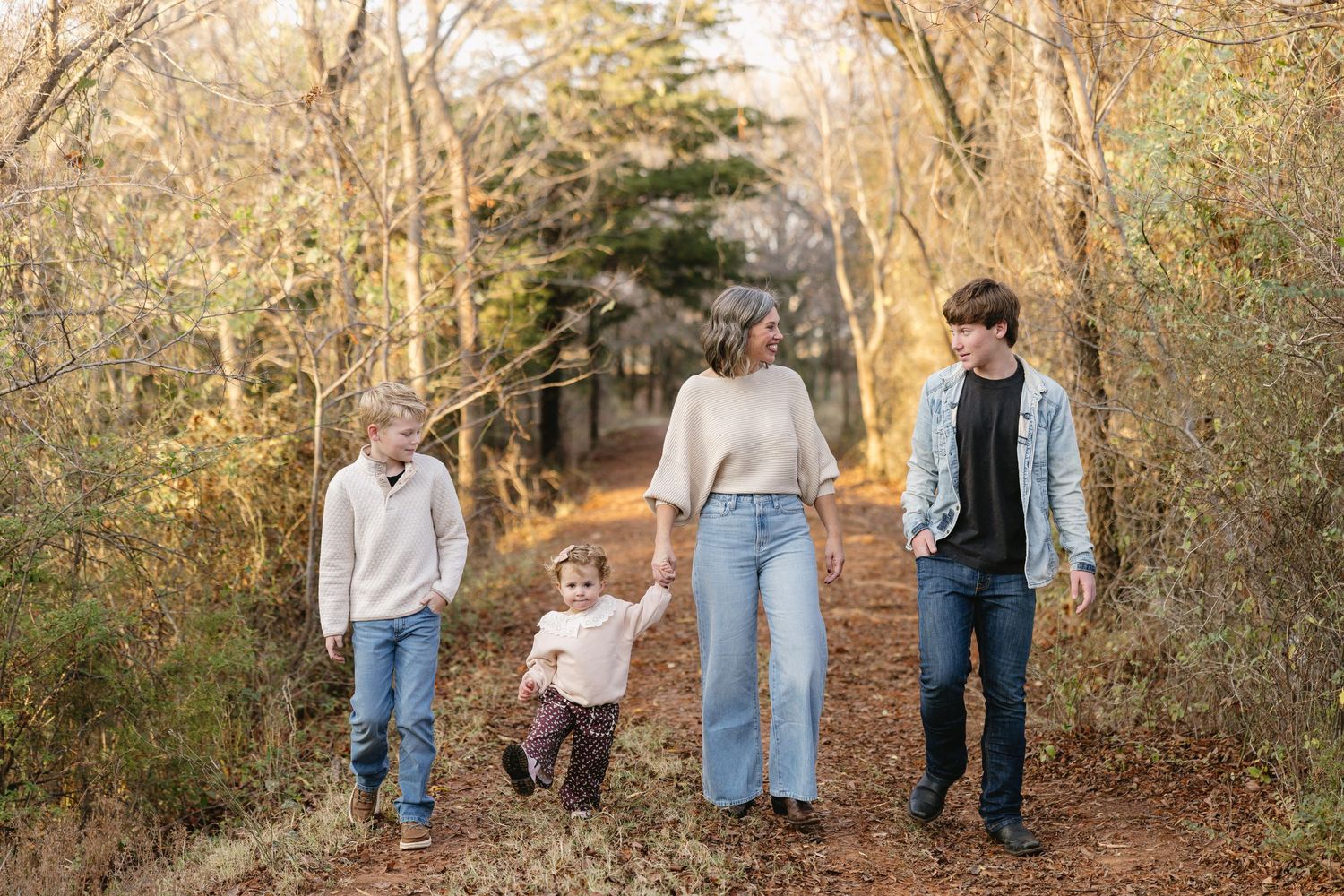 Family of four walking on a wooded trail in autumn, parents holding hands with young children amid golden foliage.