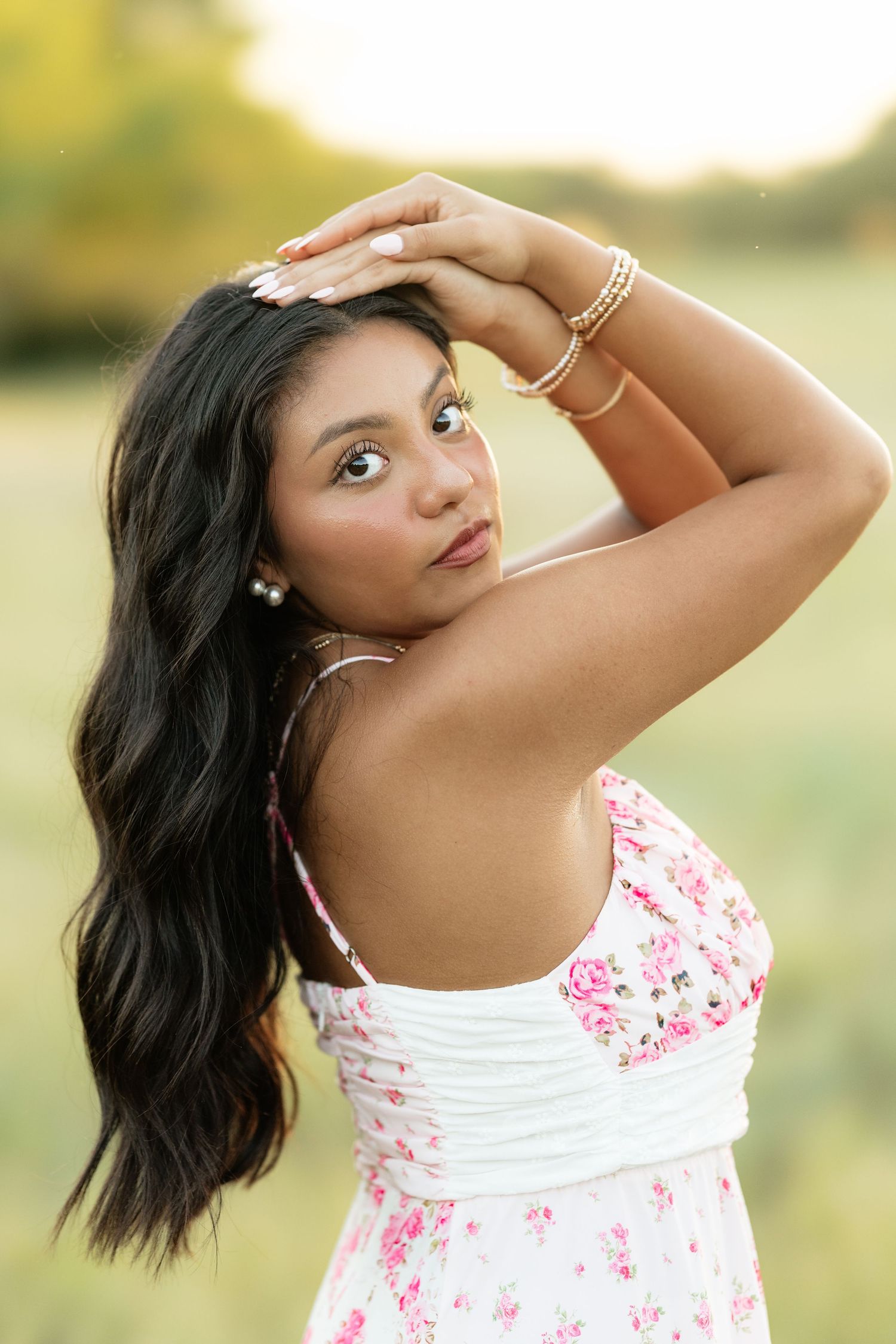 Young woman in floral dress poses outdoors in golden hour light, wearing gold bracelets and pearl earrings.