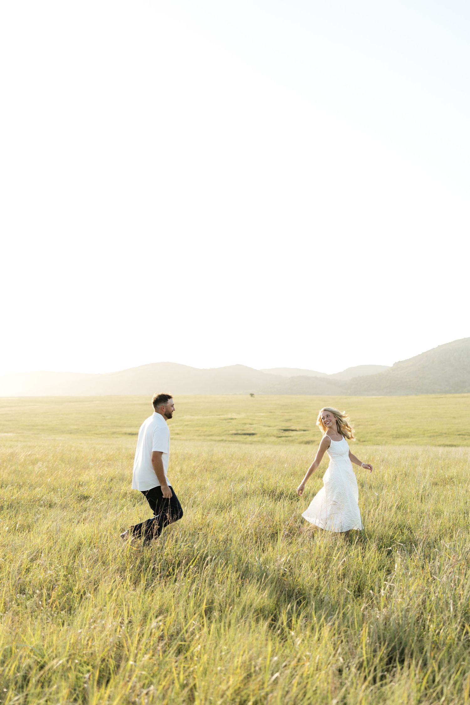 A couple walks through a sunlit green meadow, the woman in a white dress and man in white shirt, with mountains in the background.