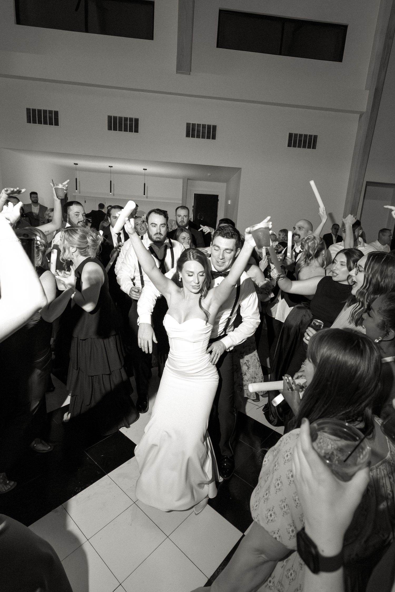 Black and white photo of bride and groom dancing with guests at a lively wedding reception celebration.