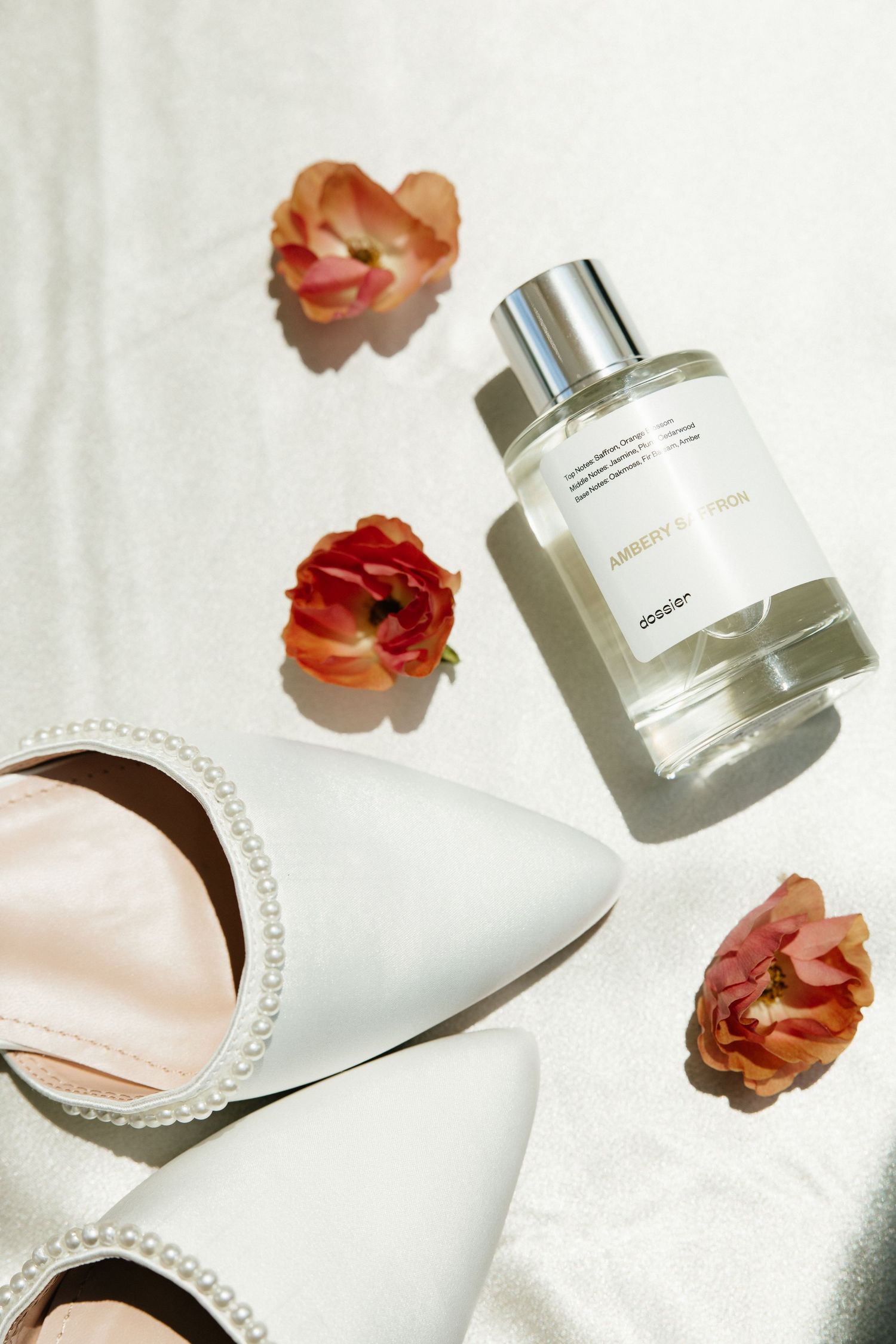 Bridal flat lay featuring pearl-trimmed white heels, Dôsér perfume bottle, and pink flowers on white fabric.