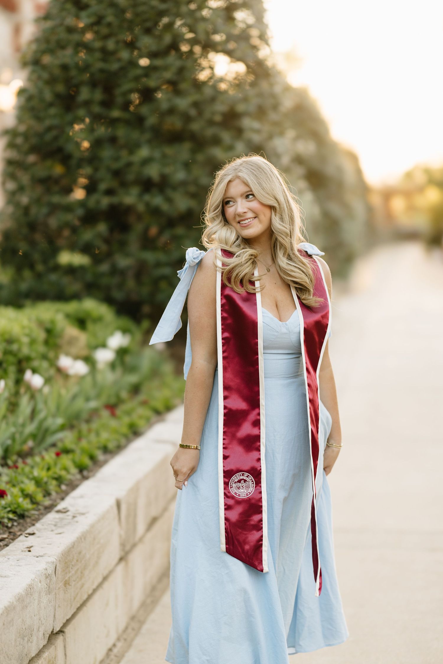 Smiling graduate in light blue dress with maroon graduation stole outdoors.