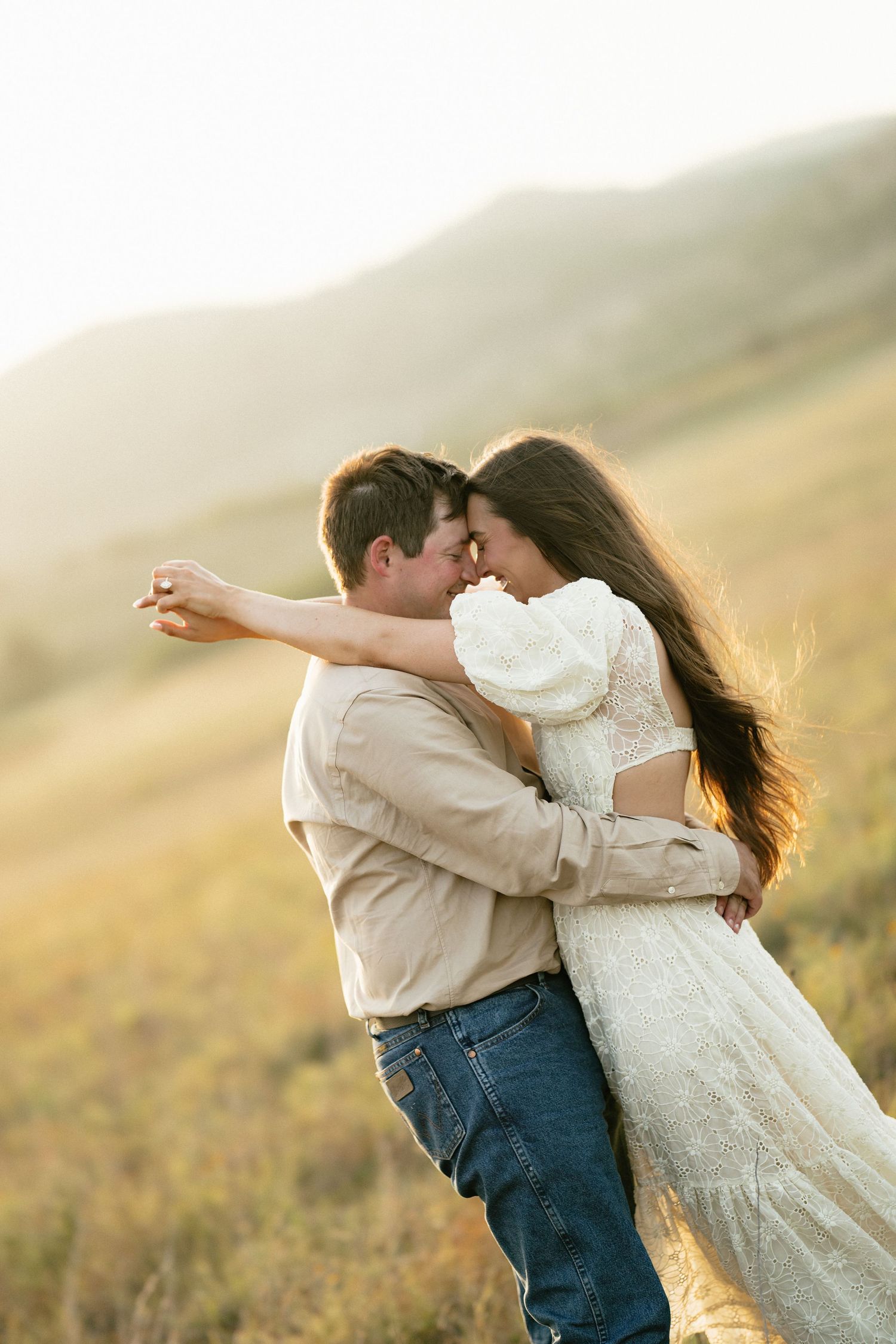 A couple sharing a romantic embrace in a sunlit open field with mountains in the background.