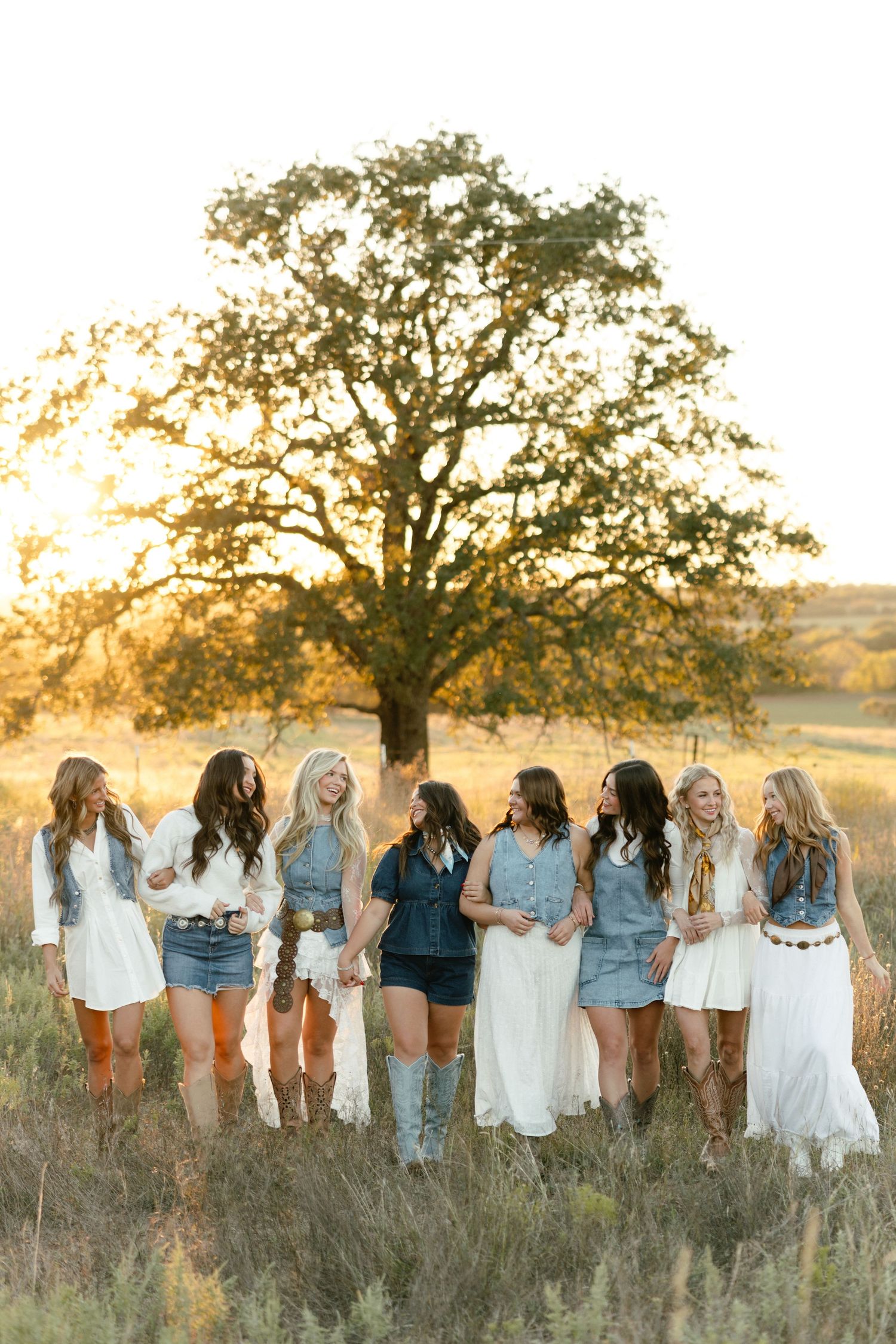 Group of bridesmaids in denim and white outfits with cowboy boots posing at golden hour near oak tree.
